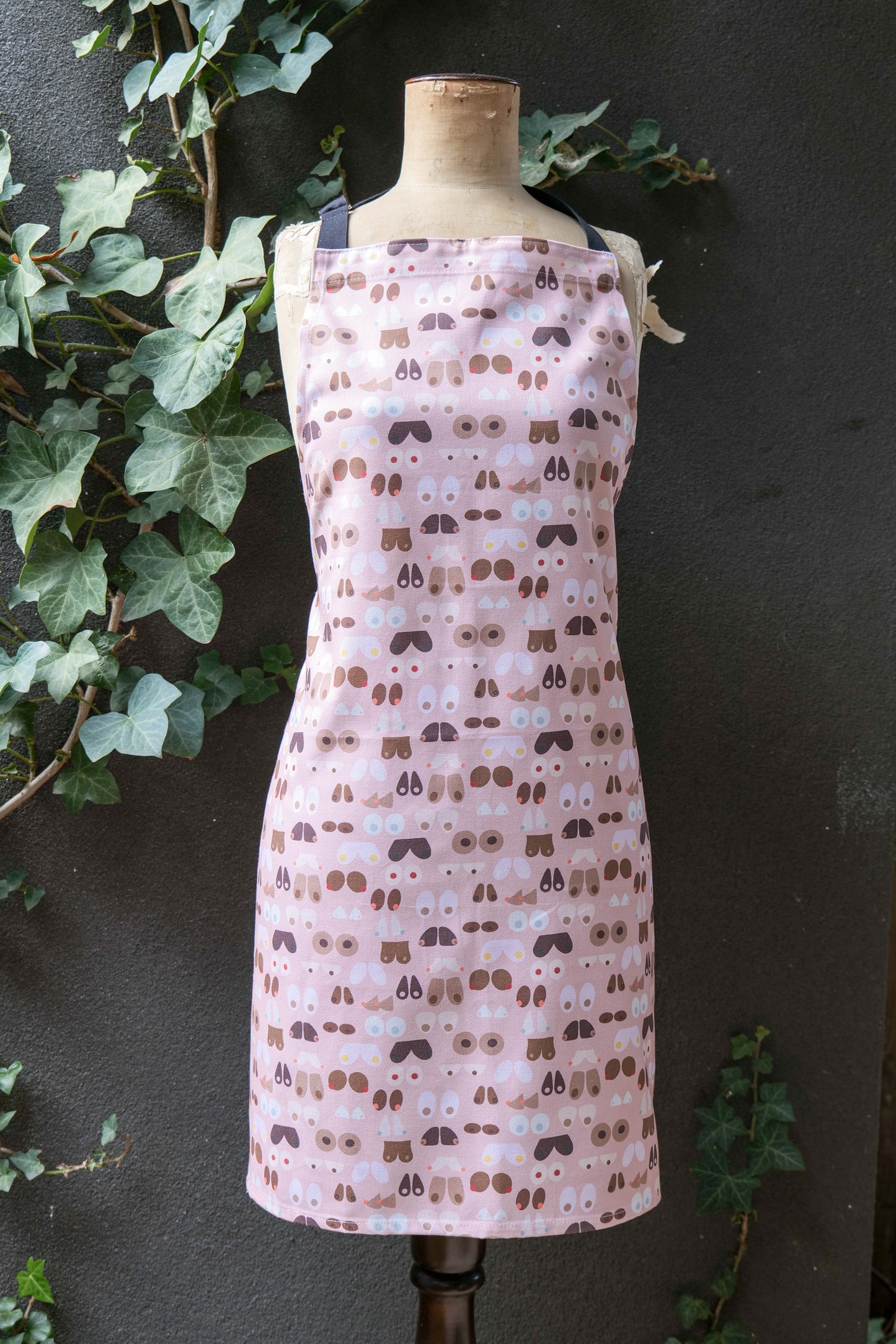 Pale pink apron featuring illustrated breasts in varied shapes, sizes and skin tones, modelled on a mannequin in front of a dark background; the apron has an adjustable neck strap, extended waist ties and a front pocket; generous fit, handmade in Australia from cotton canvas and machine washable.