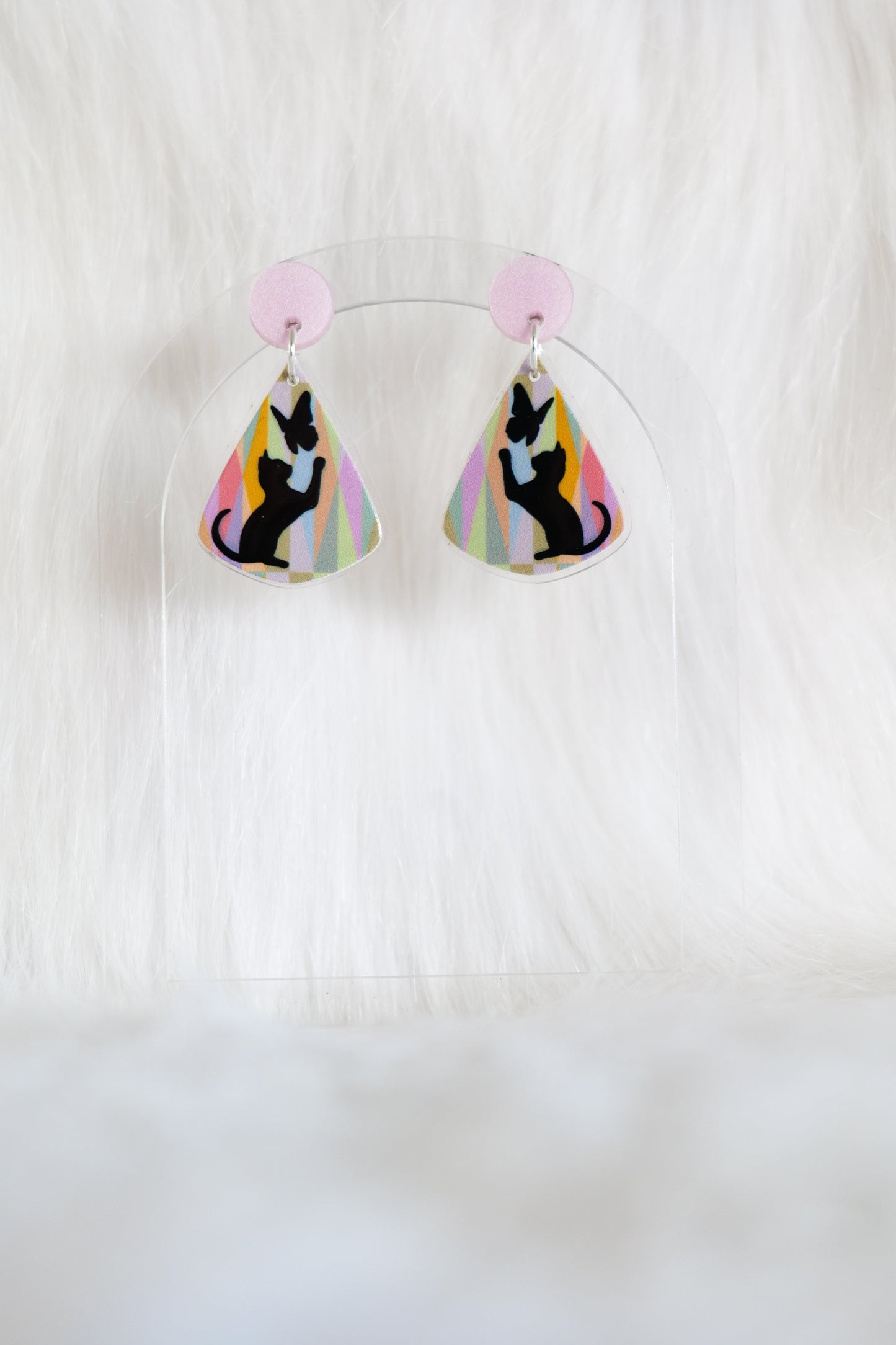 Teardrop shaped earrings featuring black cat silhouette reaching toward a butterfly on a pastel geometric background with a pale pink stud top, displayed on a clear stand against a white background