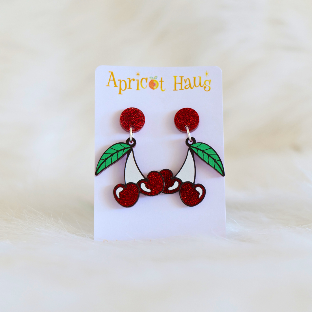 Earrings in the shape of a pair of cherries with a large green leaf, printed on red glitter acrylic with a red glitter stud at the top, lightweight with accessible large backs and stainless steel posts, designed and made in Australia