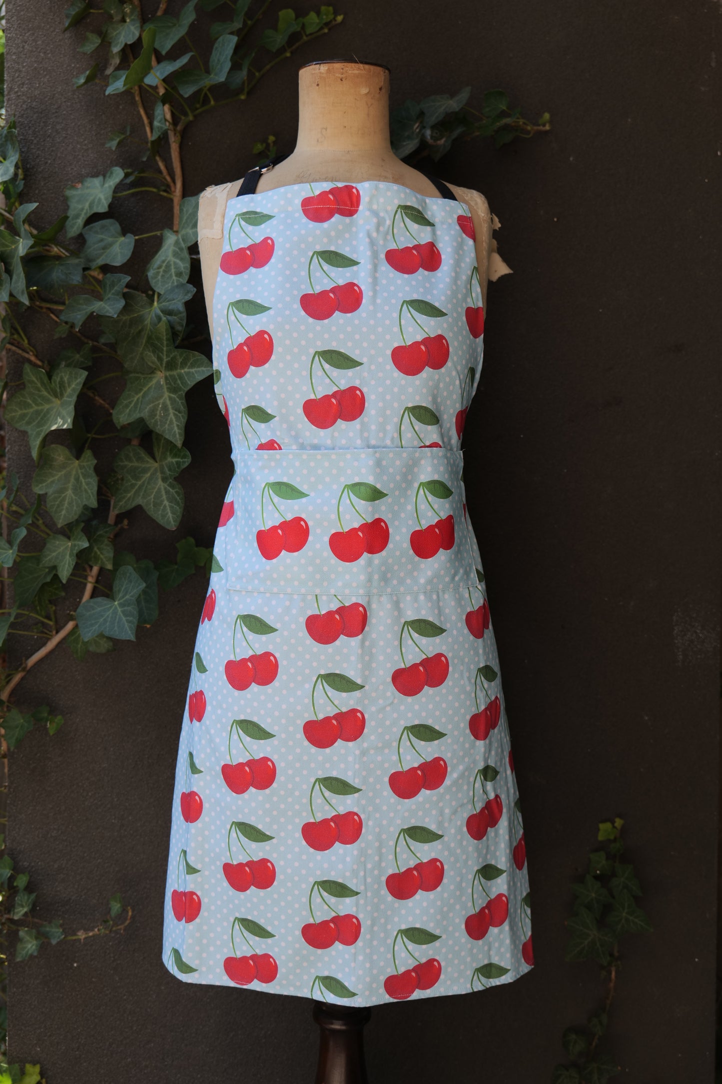 Cherries on Blue apron has a pale blue background with white polka dots and pairs of red cherries printed all over, modelled on a mannequin in front of a dark background; made of heavyweight cotton with extended waist ties and front pocket, made in Australia