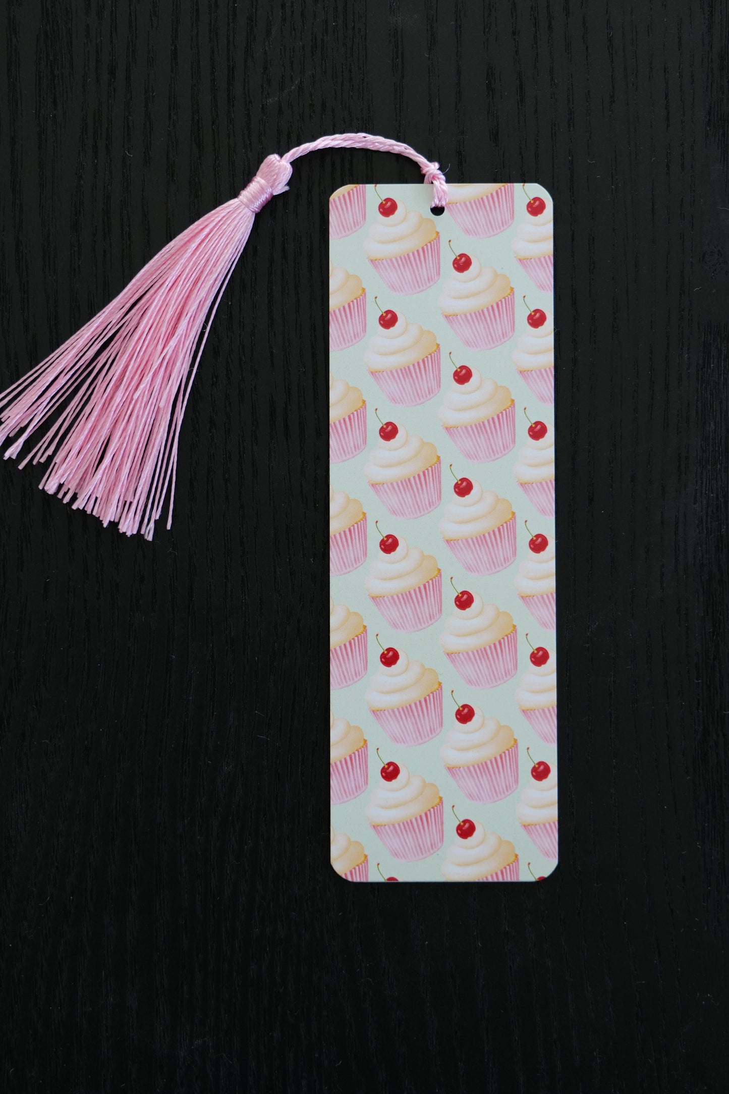Bookmark featuring a repeating pattern of pale pink cupcakes with cherries on top with a light green background and a pale pink tassel, sitting against a black backdrop