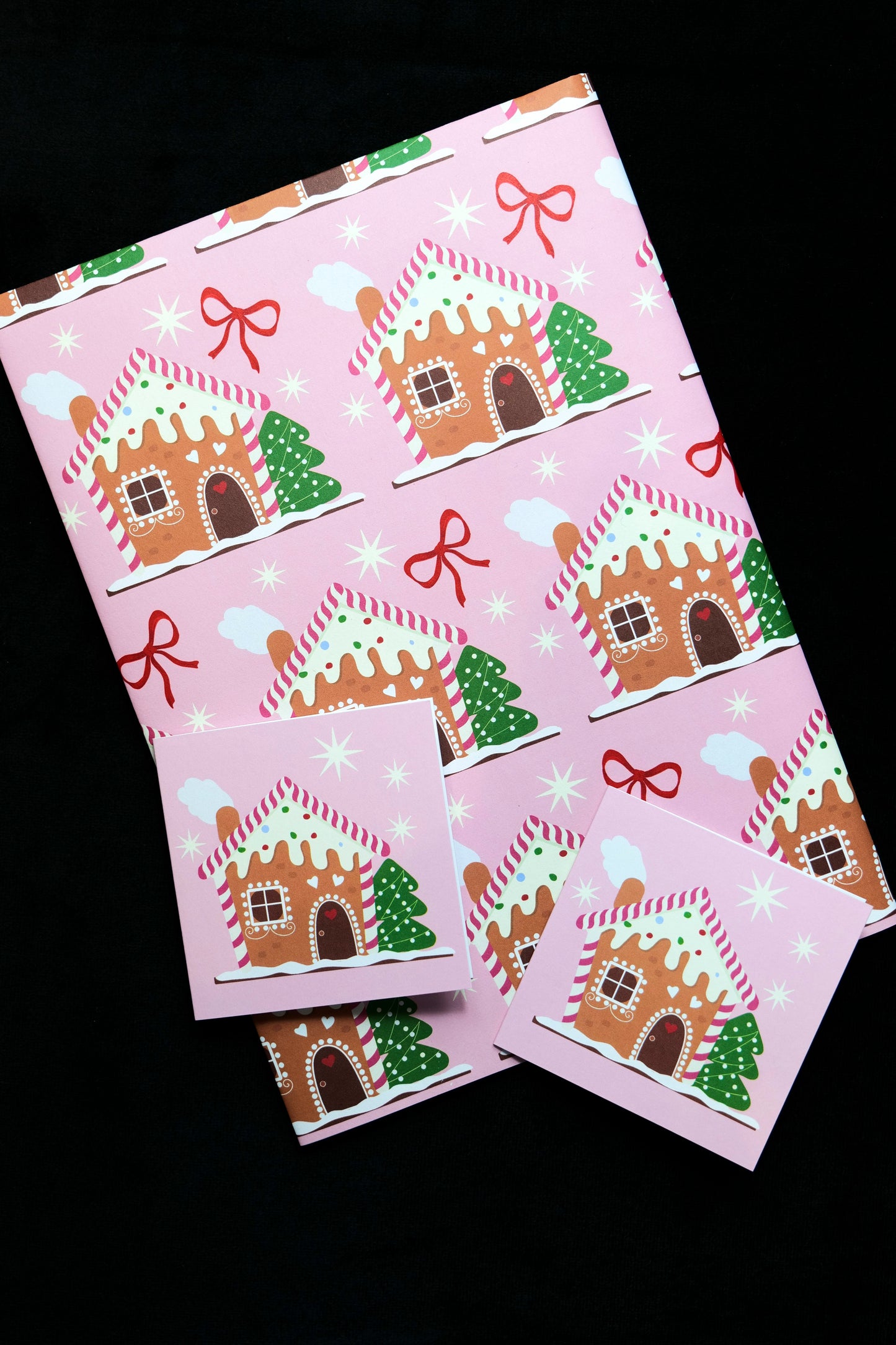 Sheet of folded gift wrap featuring gingerbread houses, stars and red bows on a pink background with two matching gift cards, displayed against a black backdrop