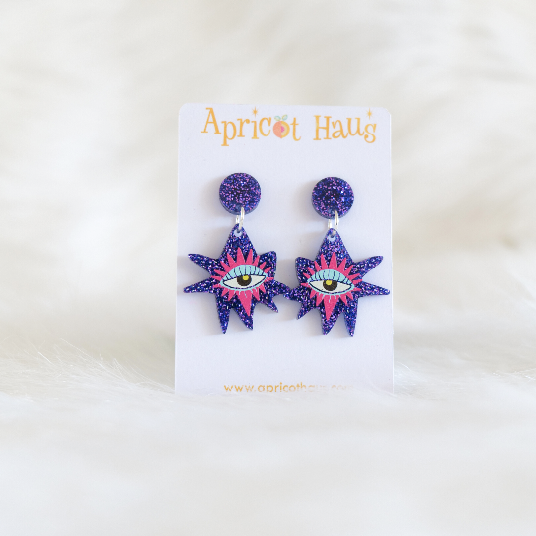 Pair of lightweight quirky star shaped earrings printed on purple glitter acrylic with a pink and blue eye in the centre, round purple glitter stud, hypoallergenic with large backs, designed and handmade in Australia, displayed on an Apricot Haus earring card