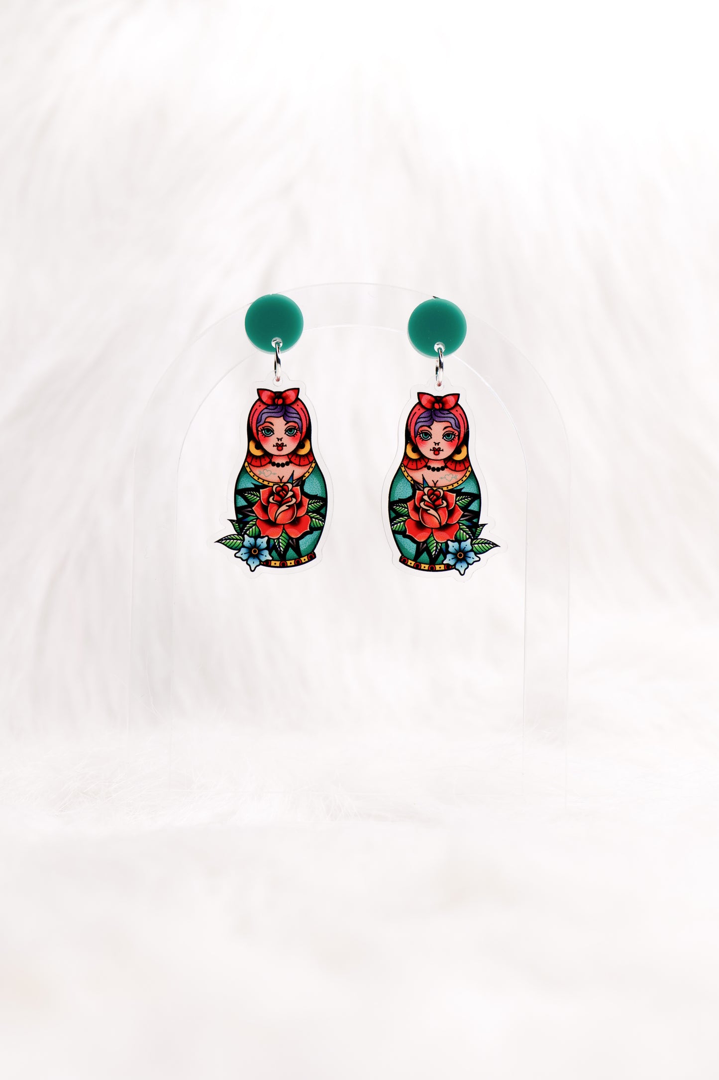 Earrings featuring colourful retro tattoo-style matryoshka dolls with a red rose and a teal stud top, displayed on a clear stand against a white background