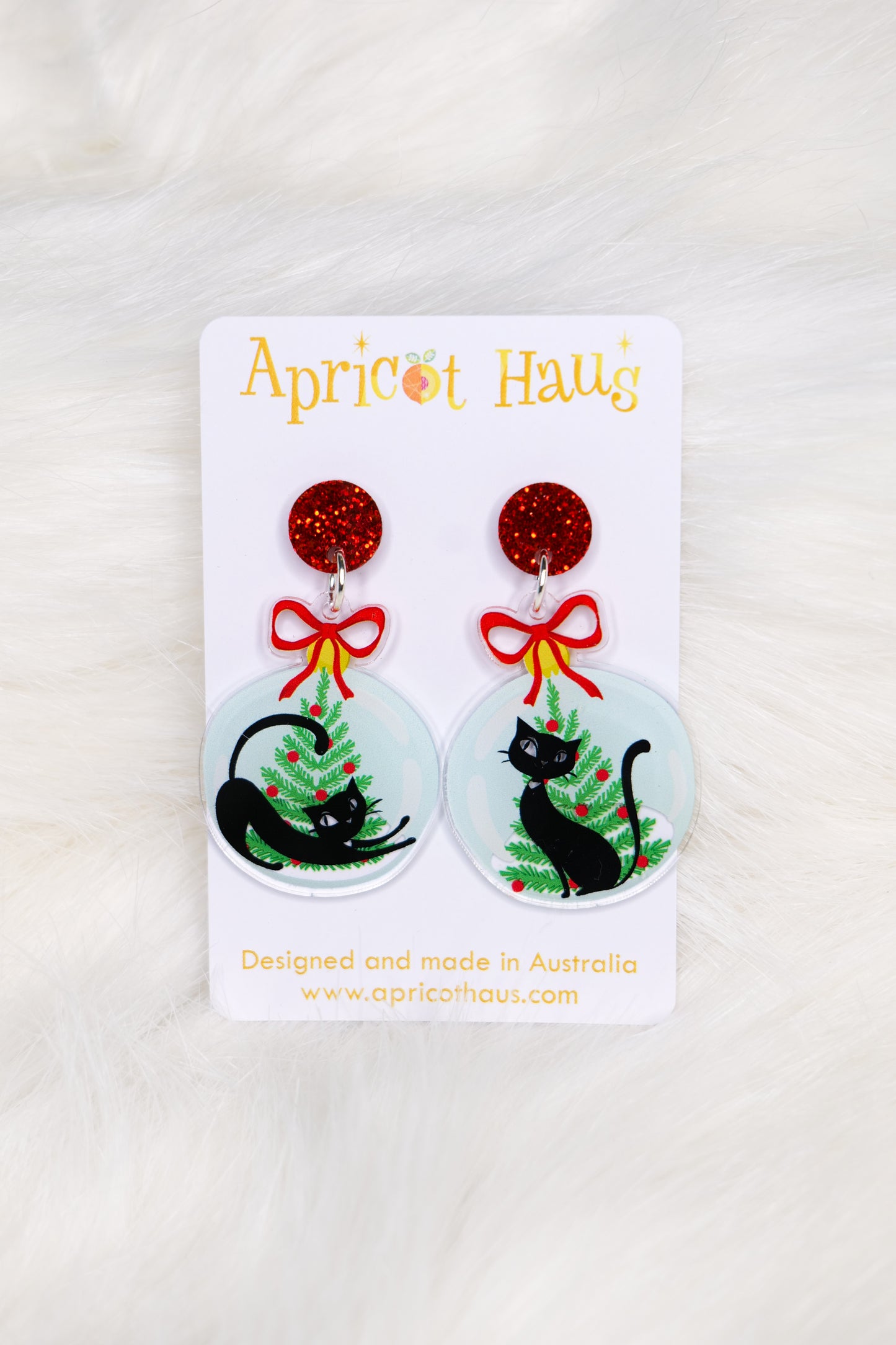 Christmas earrings in the shape of a Christmas bauble with retro-styled black cats and trees, topped with red bows and a red glitter stud; displayed on an Apricot Haus earring card on a white background