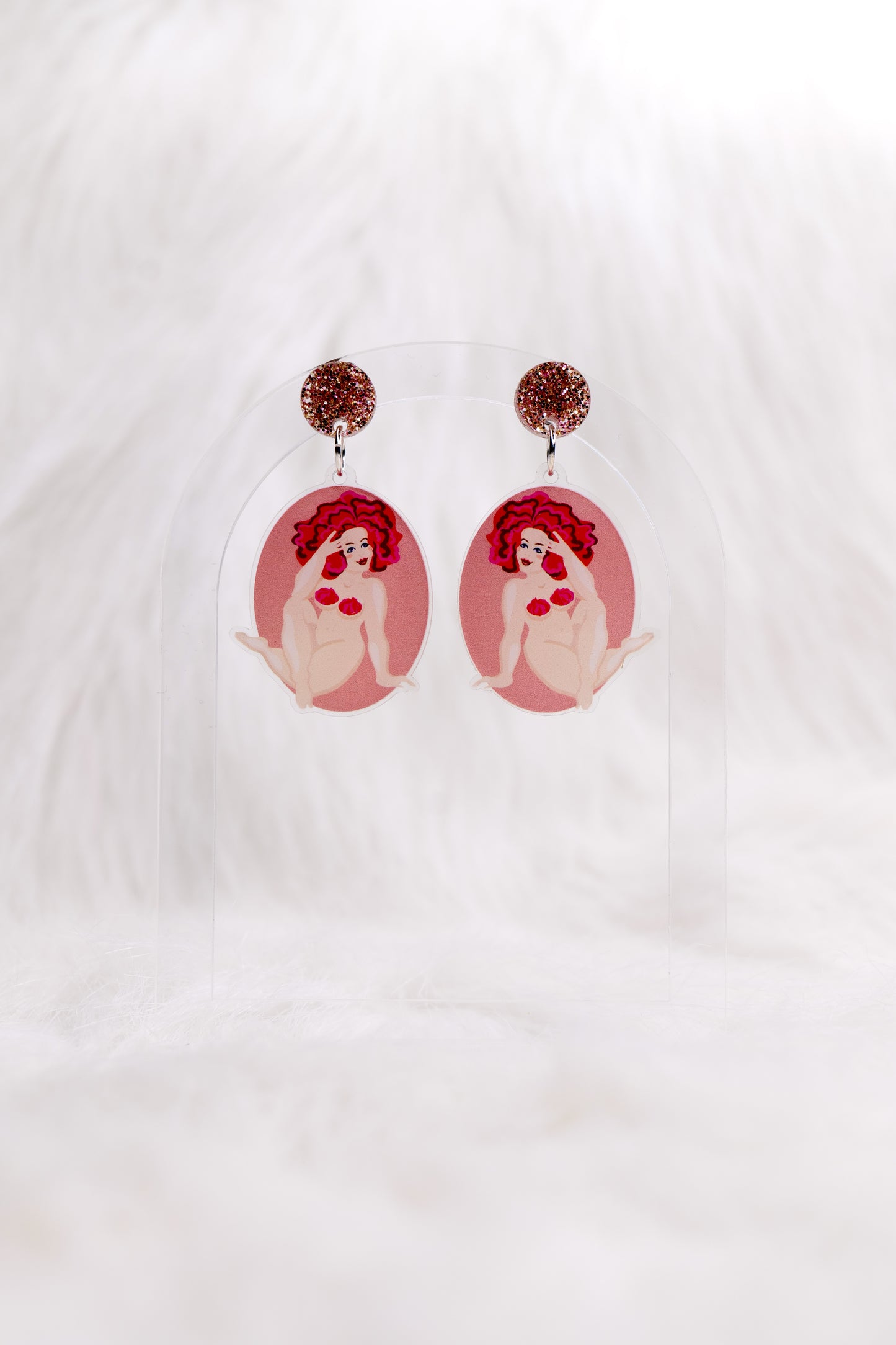 Pair of lightweight acrylic earrings in an oval shape, featuring an illustration of a nude woman with red hair and strategically placed red frosting on a pink background, earrings have stainless steel posts and large accessible back, displayed against a white backdrop