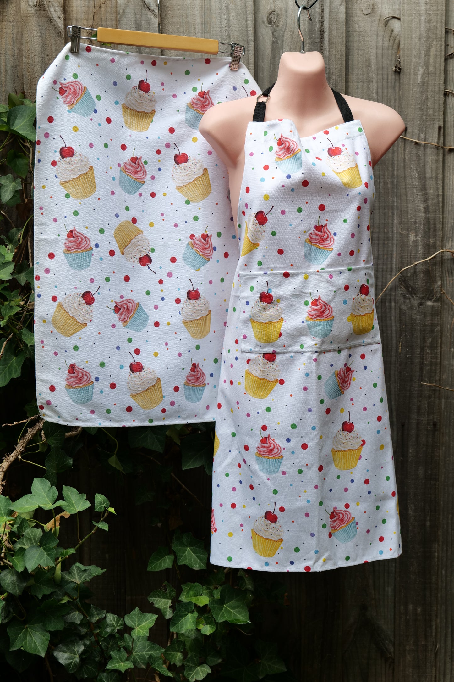 Apron and tea towel with white background covered in colorful cupcake illustrations and polka dots, both hanging side by side on a wooden fence. Apron features adjustable neck and long waist ties with a front pocket, both items are made in Australia from premium cotton