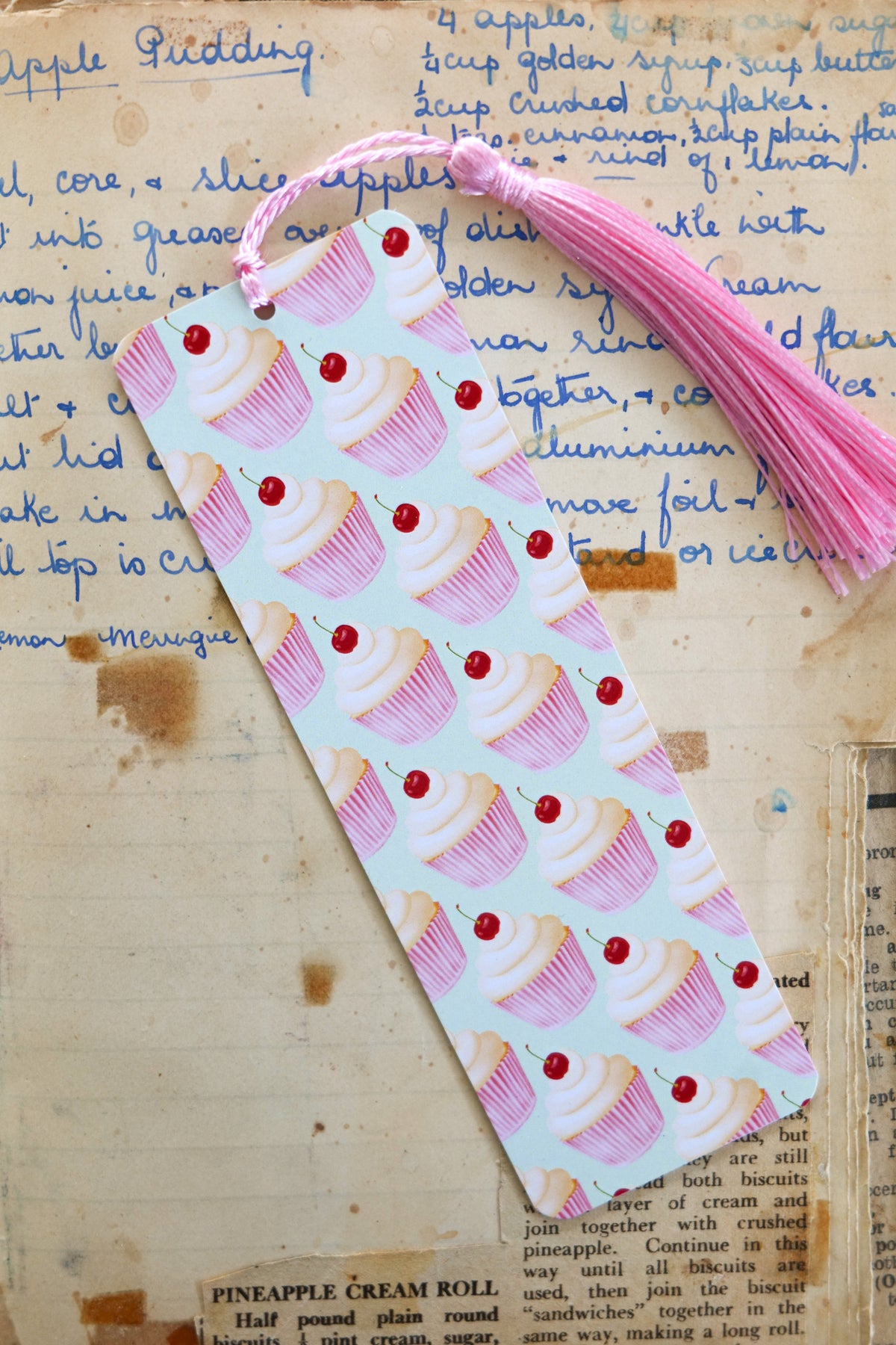 Picture of a bookmark with pink cupcakes in a grid pattern on a pale green background and a pink tassel, sitting on a handwritten recipe