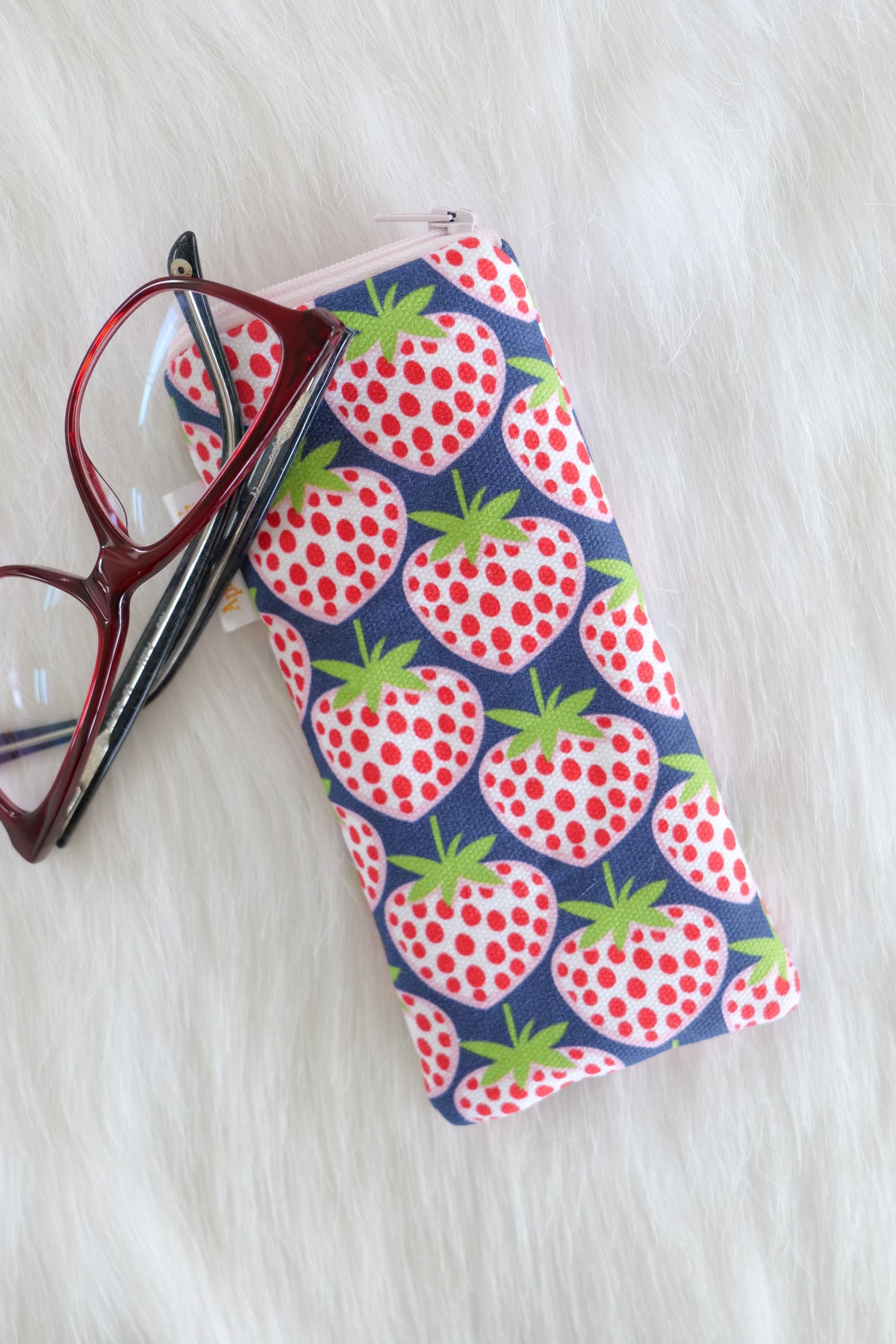 Blue strawberries glasses case featuring red and white strawberries on a navy background, displayed on a white background alongside a pair of eyeglasses. 