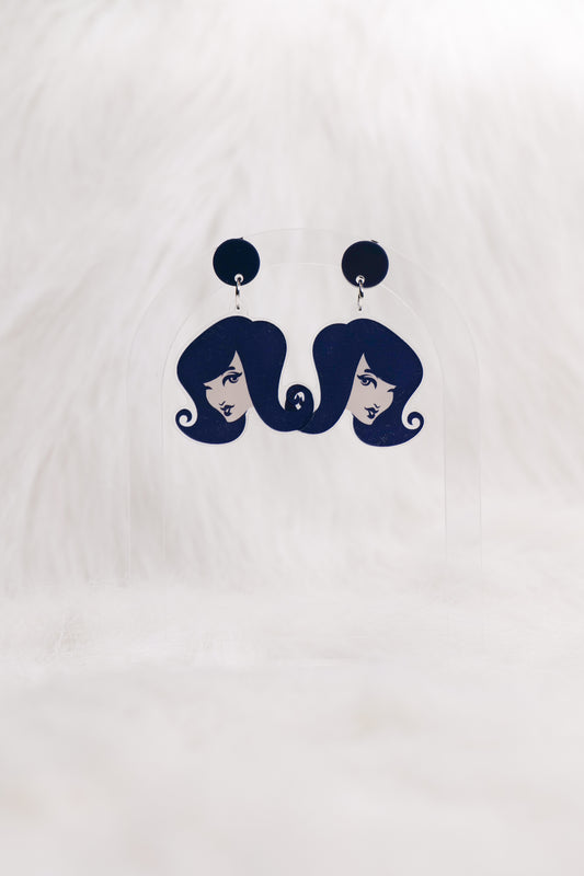 Earrings featuring graphic style girls with 60s style hair with circular charcoal-coloured studs, displayed on a clear stand against a white background