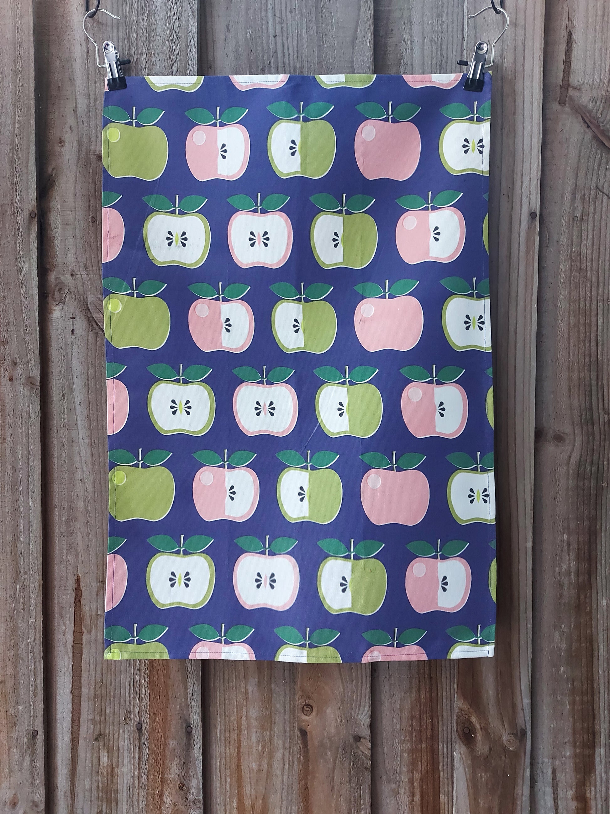 Cotton tea towel with a retro 70s‑inspired pattern of stylised apples in green, pink and cream on a navy background, displayed against a wooden fence