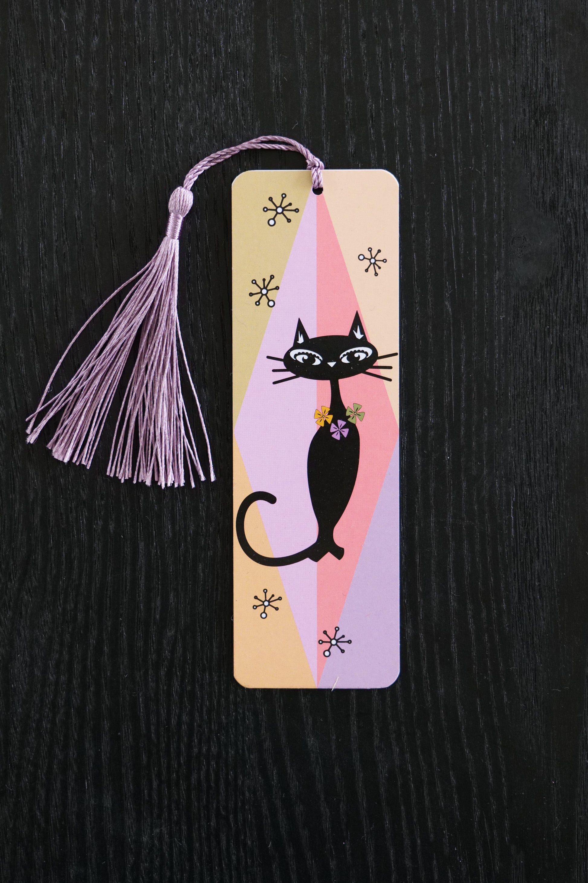 Bookmark with a black 50s style cat against a geometric background in pink, mauve and khaki; a mauve tassel against a black backdrop