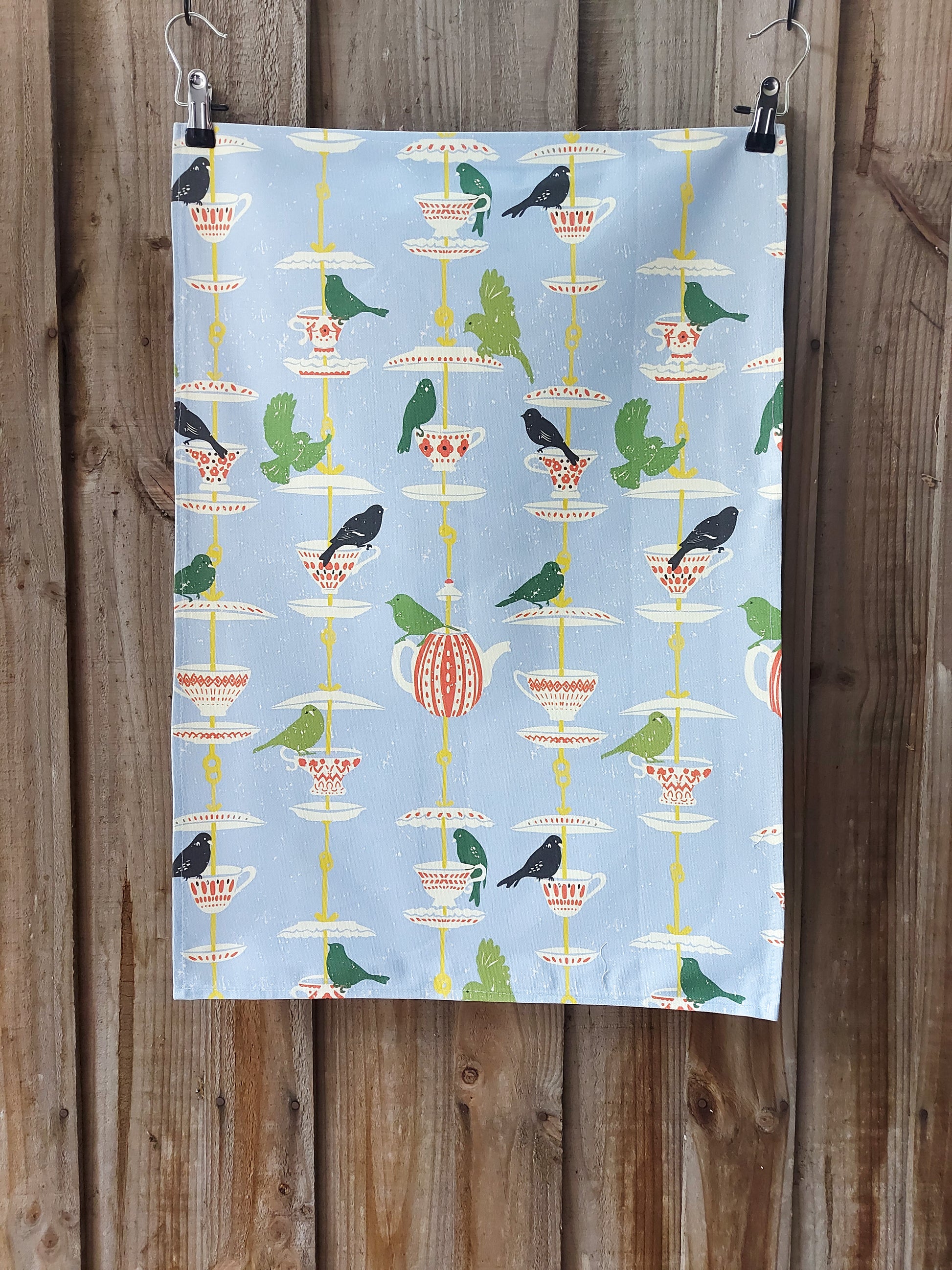Cotton tea towel featuring a Scandi/folk‑inspired pattern of green and black birds perched on stacked teacups and pots on a pale blue background; displayed against a wooden fence