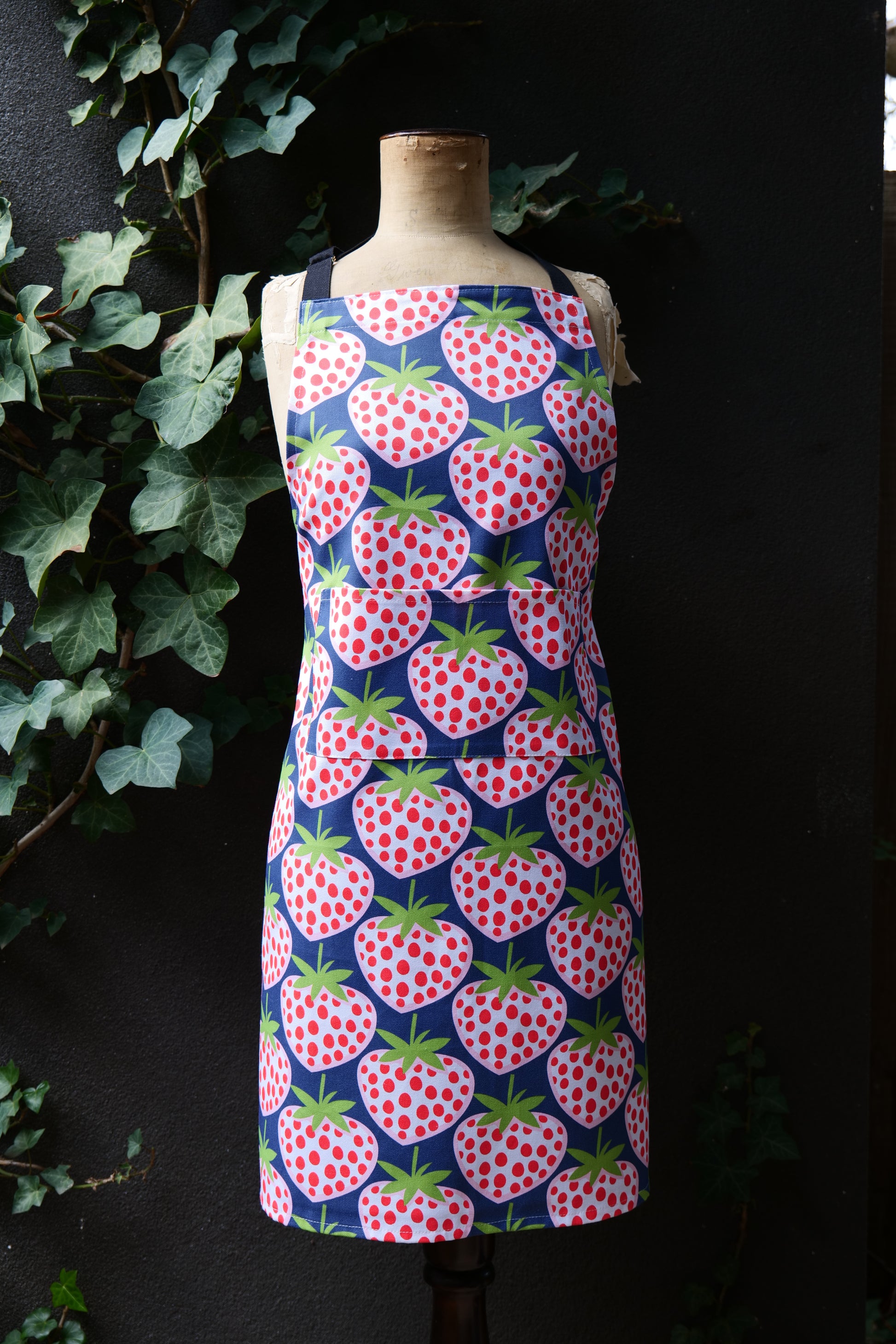 Bib-style apron featuring large red and white strawberries on a dark blue background, displayed on a mannequin against a dark backdrop