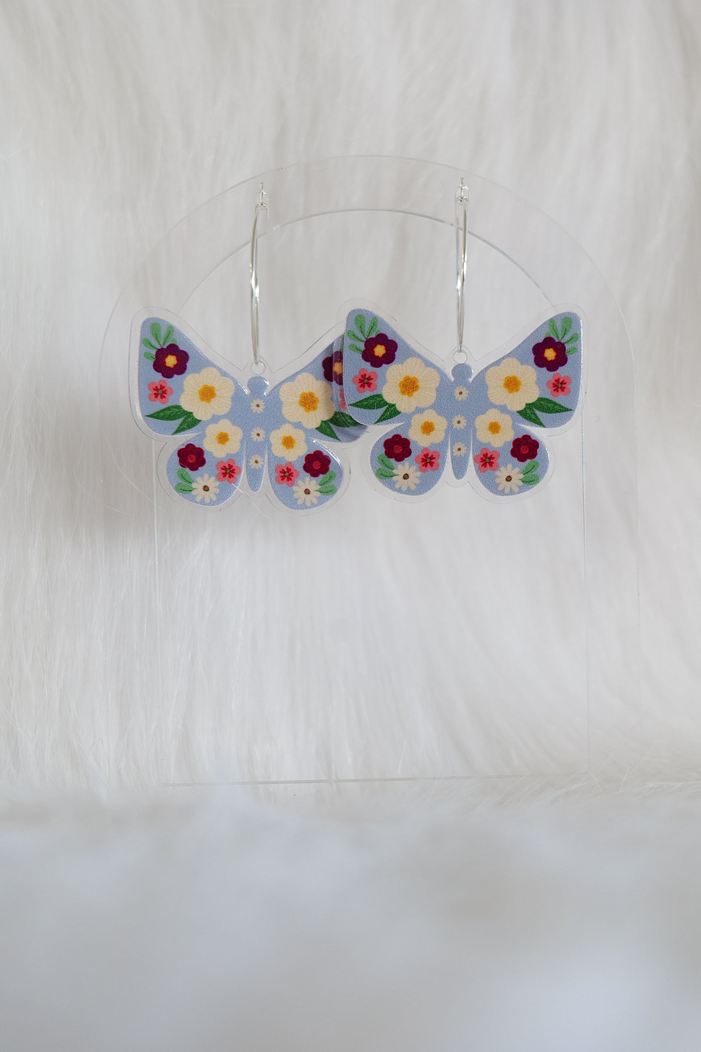 Butterfly shaped earrings with a pale blue background, covered with deep red, cream and pink flowers; printed on lightweight acrylic with stainless steel hoops, displayed against a white background
