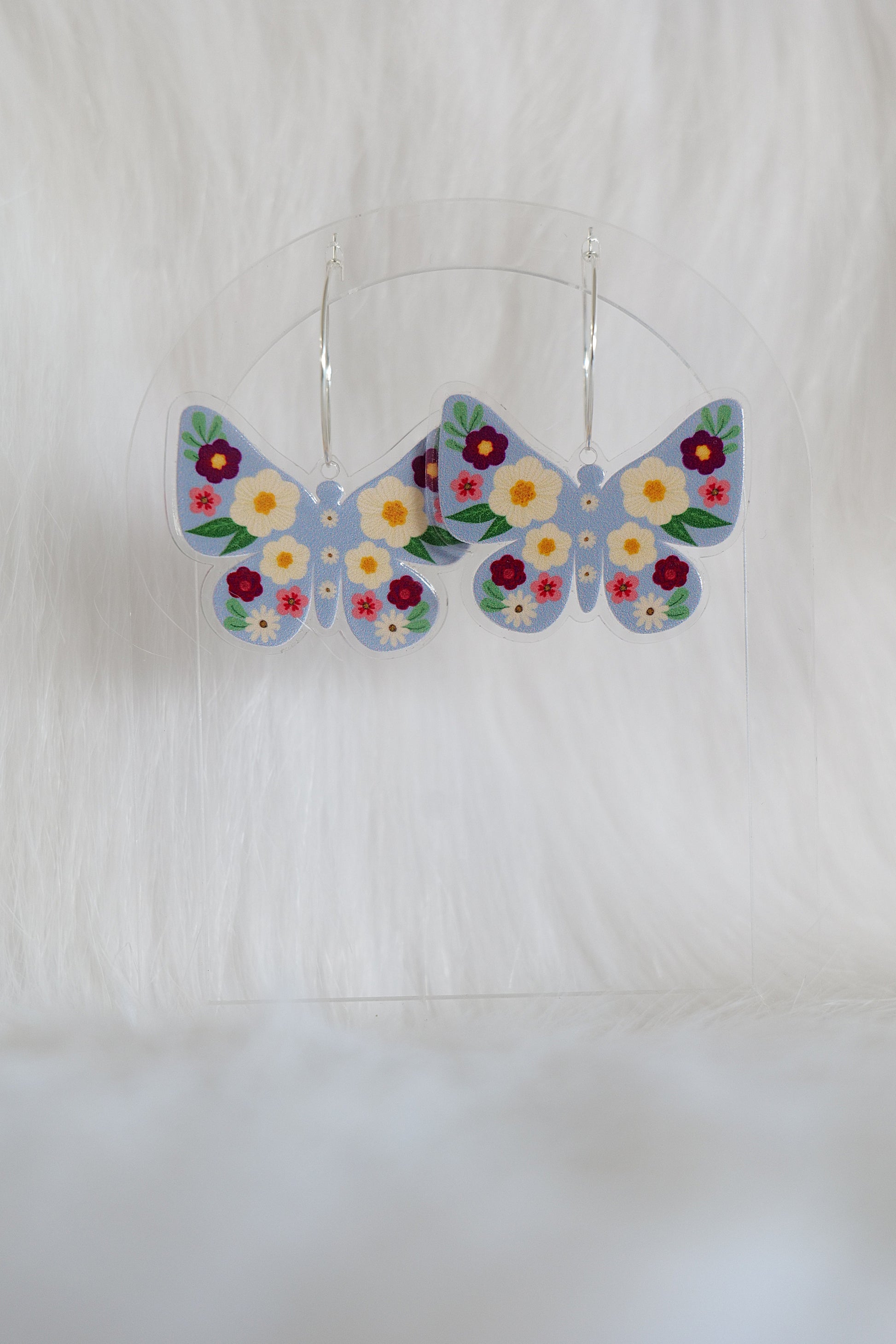 Butterfly shaped earrings with a pale blue background, covered with deep red, cream and pink flowers; printed on lightweight acrylic with stainless steel hoops, displayed against a white background