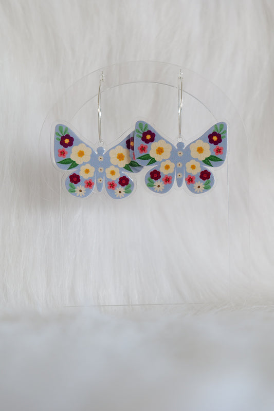 Butterfly shaped earrings with a pale blue background, covered with deep red, cream and pink flowers; printed on lightweight acrylic with stainless steel hoops, displayed against a white background