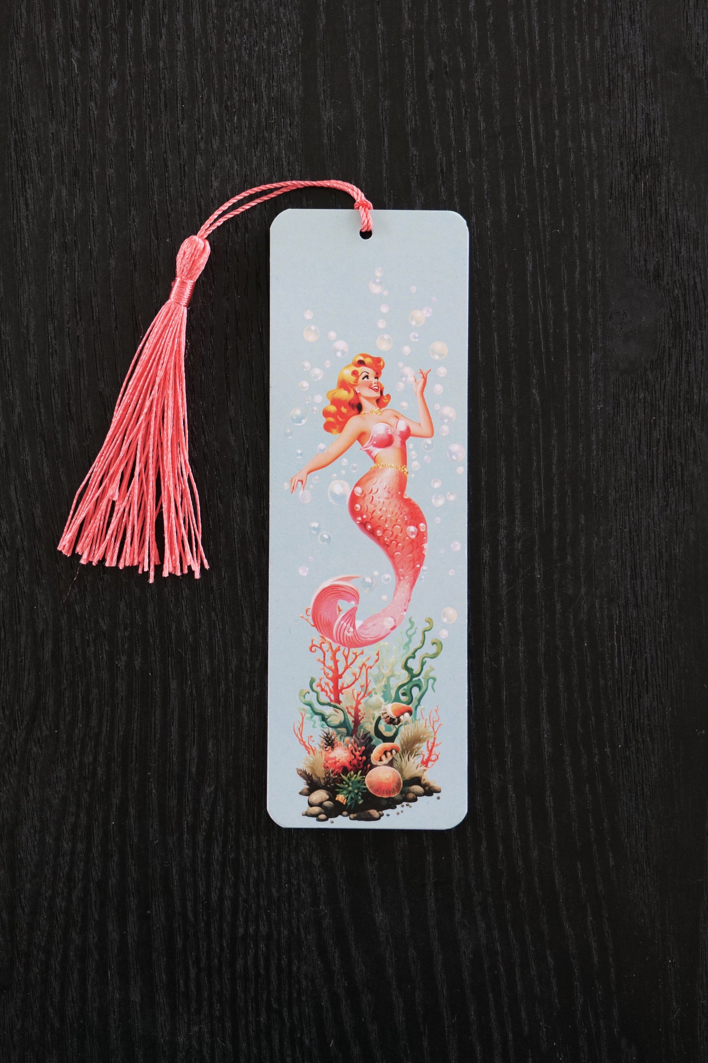 Bookmark featuring a coral coloured 50s style mermaid with seaweed and bubbles and a coral colour tassel, sitting against a black backdrop