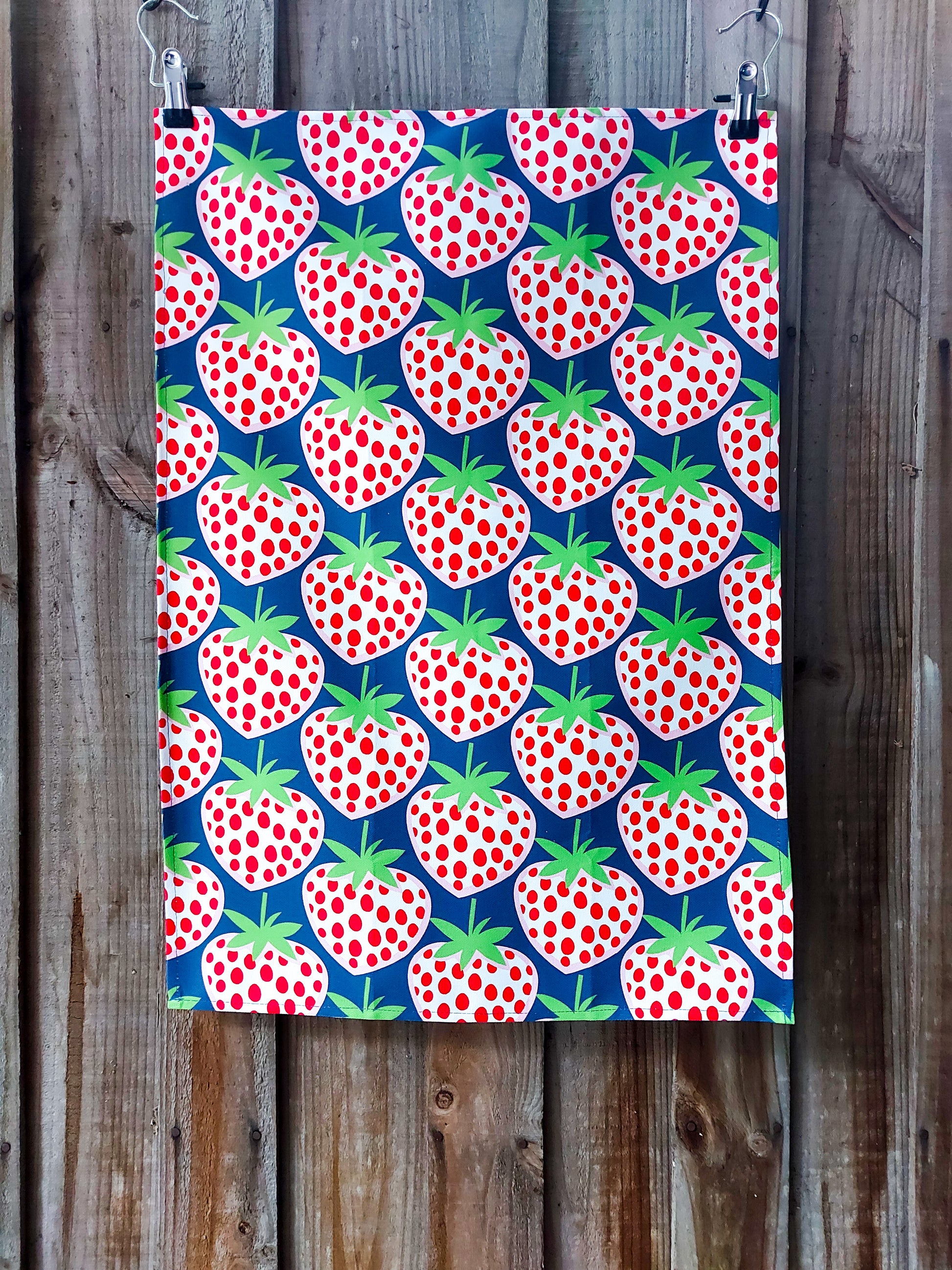 Tea towel featuring a bright all-over red and white strawberry pattern against a deep blue background; pictured against a wooden fence