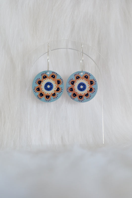 Pair of circular acrylic earrings with a blue background and brown and blue mandala-style design hanging from hoops; displayed on a clear stand against a white background
