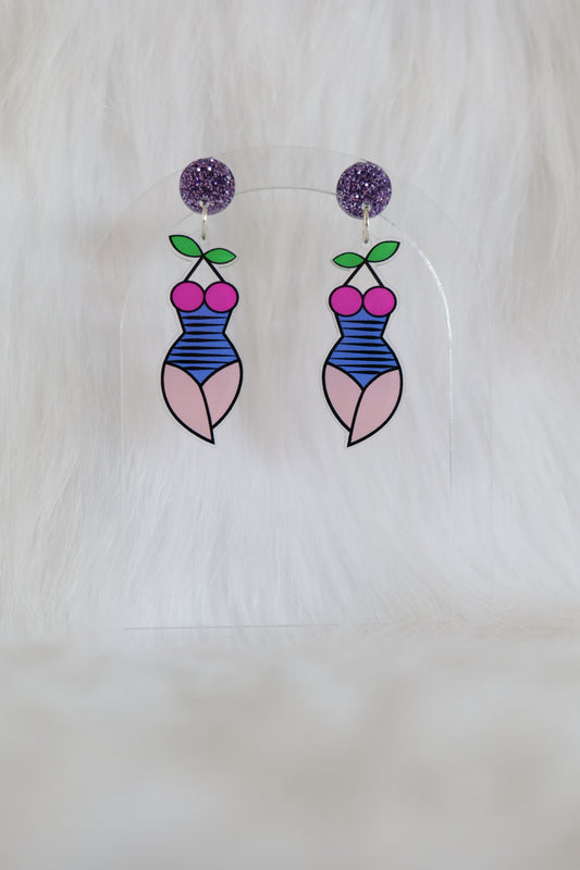 Fun pair of lightweight acrylic earrings in the shape of a female torso and thighs, wearing a striped blue swimsuit with boobs made from a pair of cherries and leaves as the halter neck, mauve glitter stud at the top, stainless steel posts and large accessible backs, displayed against a white background
