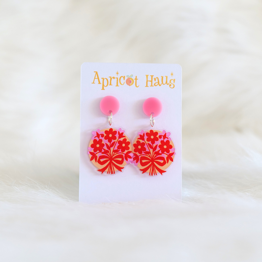 Pair of lightweight acrylic earrings printed with a flat-style illustration of red and pink flowers on a beige background, pink circular stud and stainless steel posts, handmade in Australia; displayed on an Apricot Haus earring card