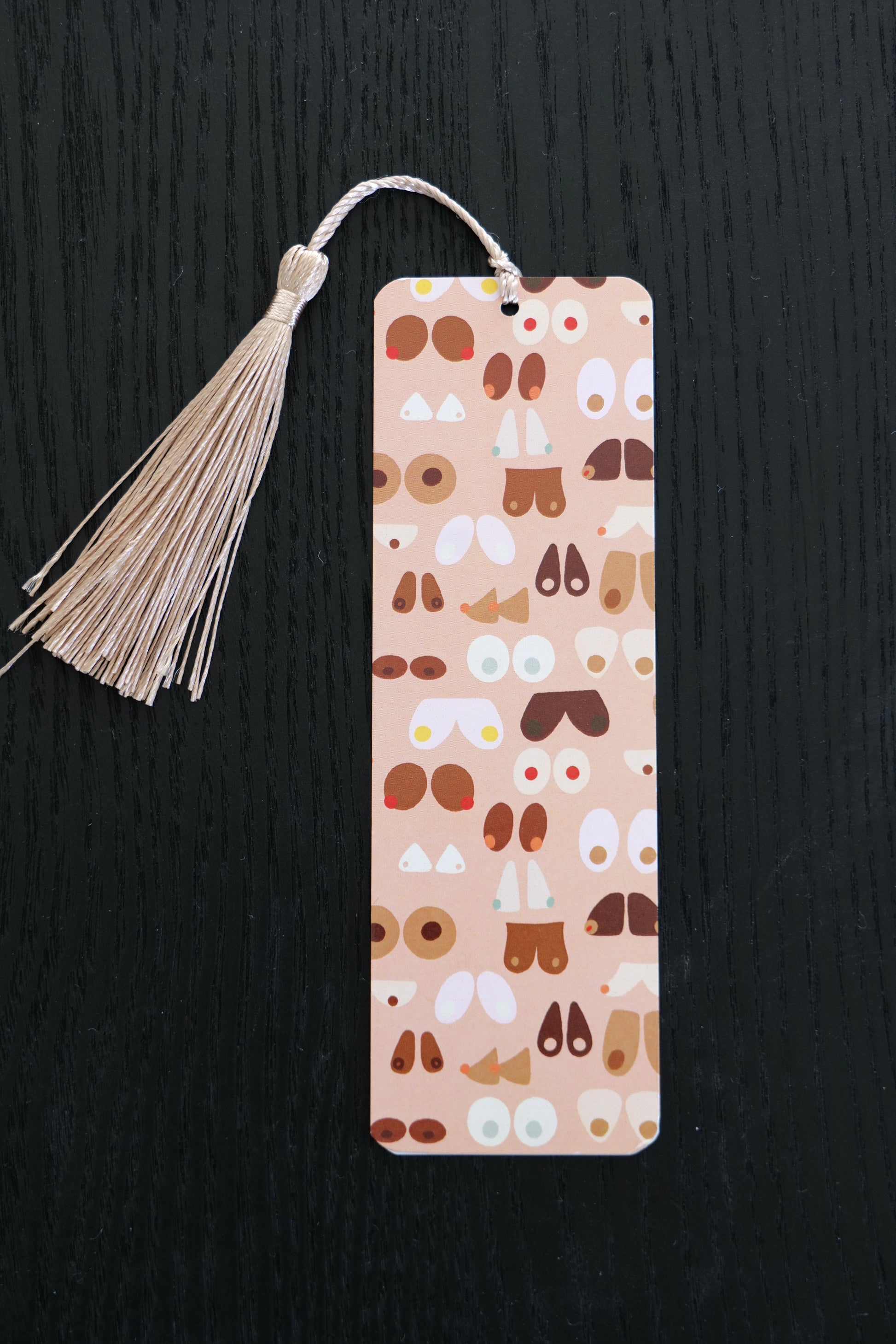 Bookmark featuring many different kinds of breasts in different colours against a pale pink background, with a beige tassel, sitting against a black backdrop