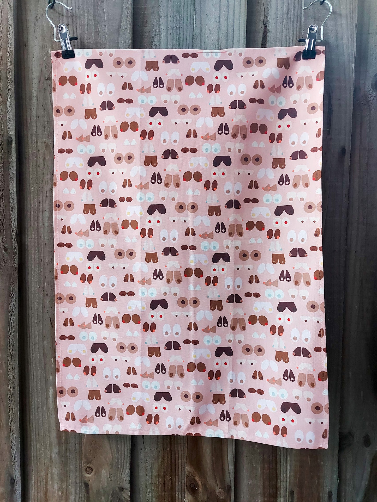 100% cotton tea towel featuring illustrated breasts in various shapes, sizes and skin tones on a pale pink background, pictured against a wooden background; certified Australian Made and Owned.