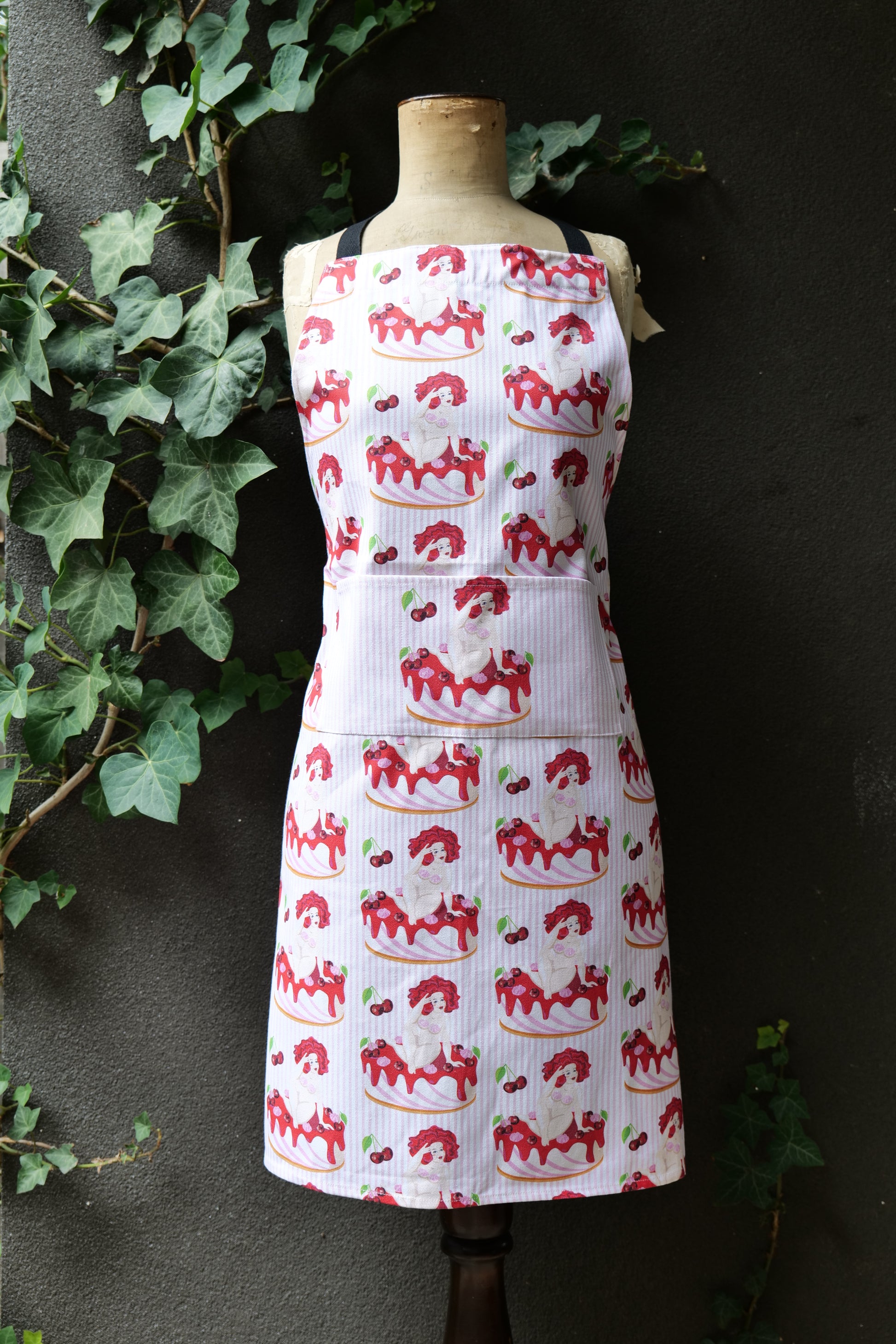Cake Nude apron features voluptuous nude women reclining on a cake on a background of pink stripes, modelled on a mannequin set against a dark background; apron has extended waist ties and adjustable neck, made from cotton canvas, machine washable and Australian made