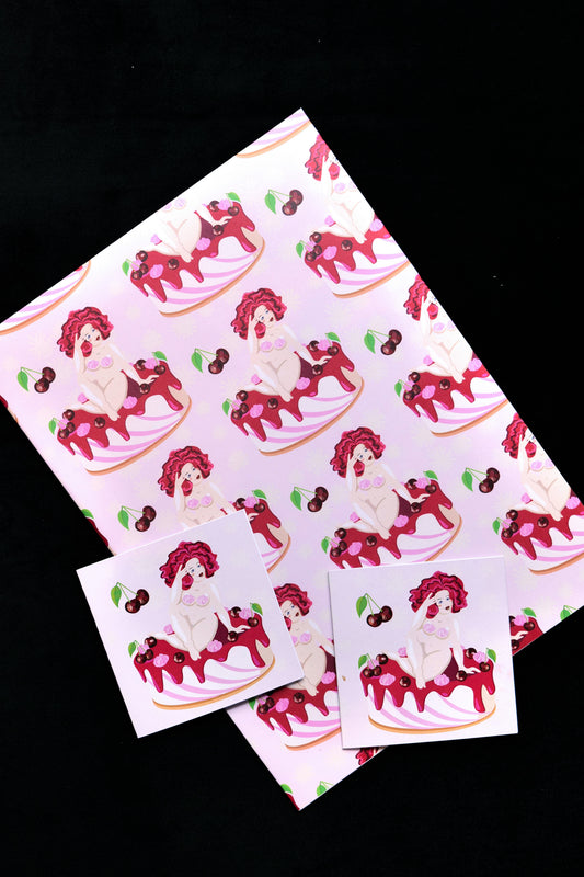A sheet of folded gift wrap with an illustration of a nude woman on a cake with cherries on a pale pink background, displayed against a black backdrop