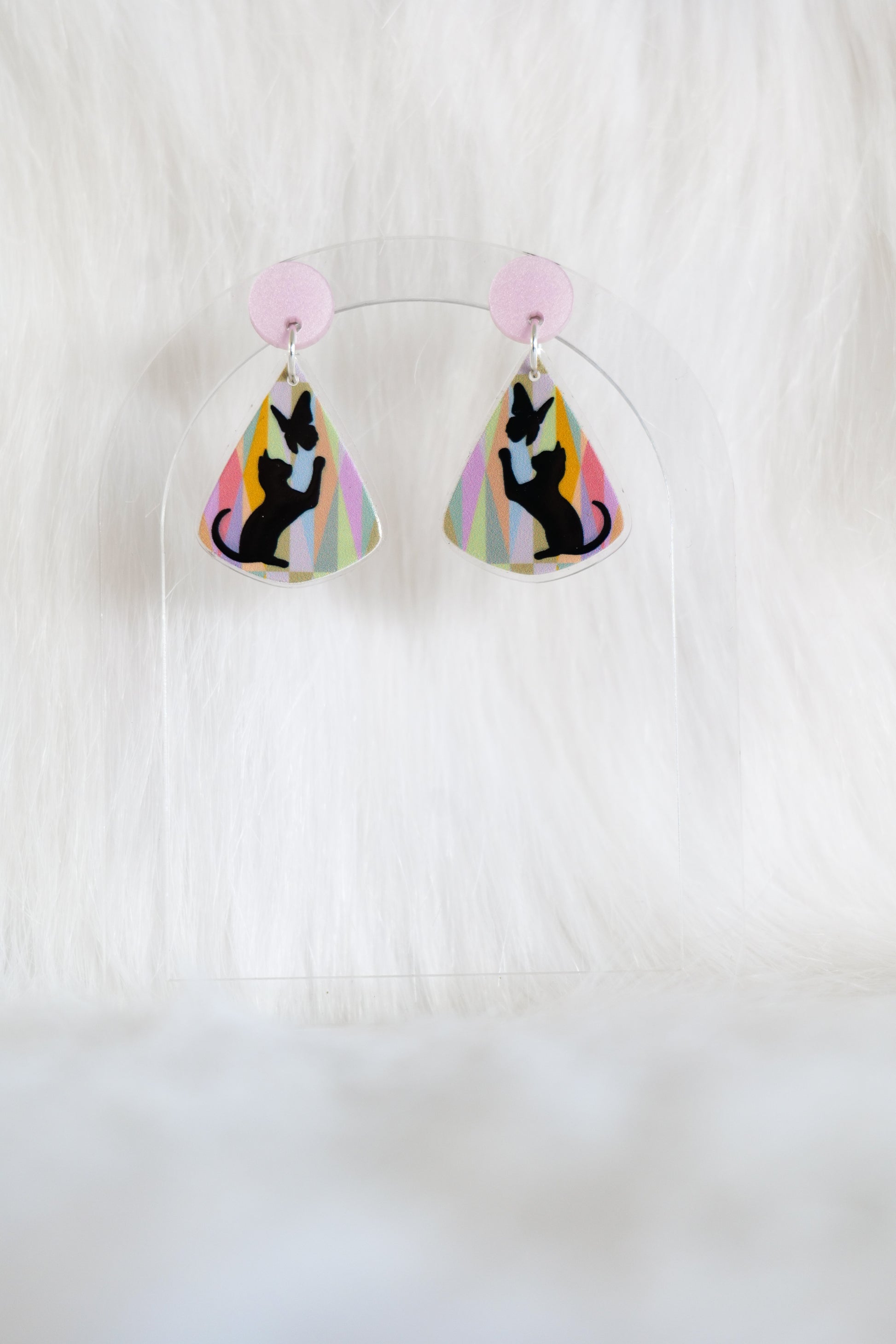 Teardrop shaped earrings featuring black cat silhouette reaching toward a butterfly on a pastel geometric background with a pale pink stud top, displayed on a clear stand against a white background