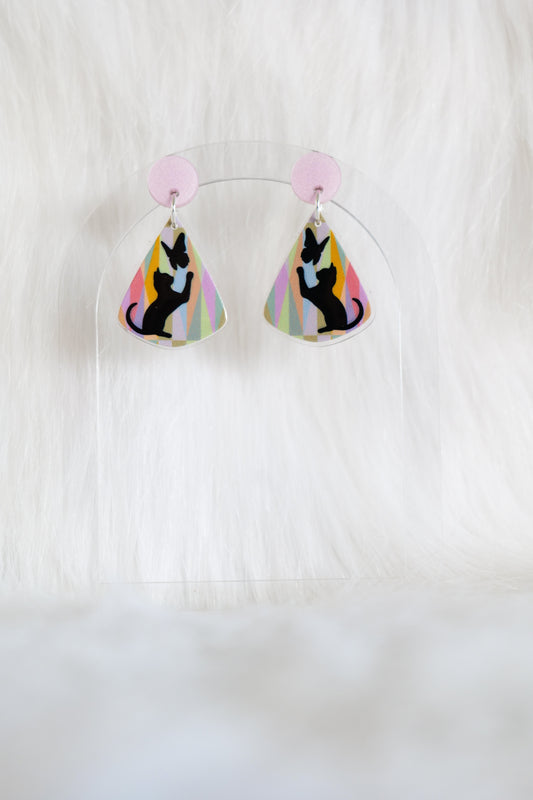 Teardrop shaped earrings featuring black cat silhouette reaching toward a butterfly on a pastel geometric background with a pale pink stud top, displayed on a clear stand against a white background