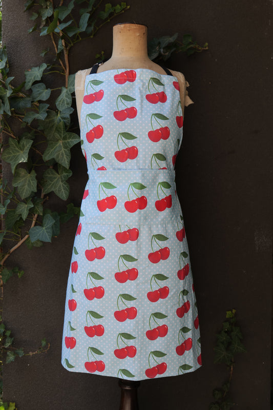 Cherries on Blue apron has a pale blue background with white polka dots and pairs of red cherries printed all over, modelled on a mannequin in front of a dark background; made of heavyweight cotton with extended waist ties and front pocket, made in Australia