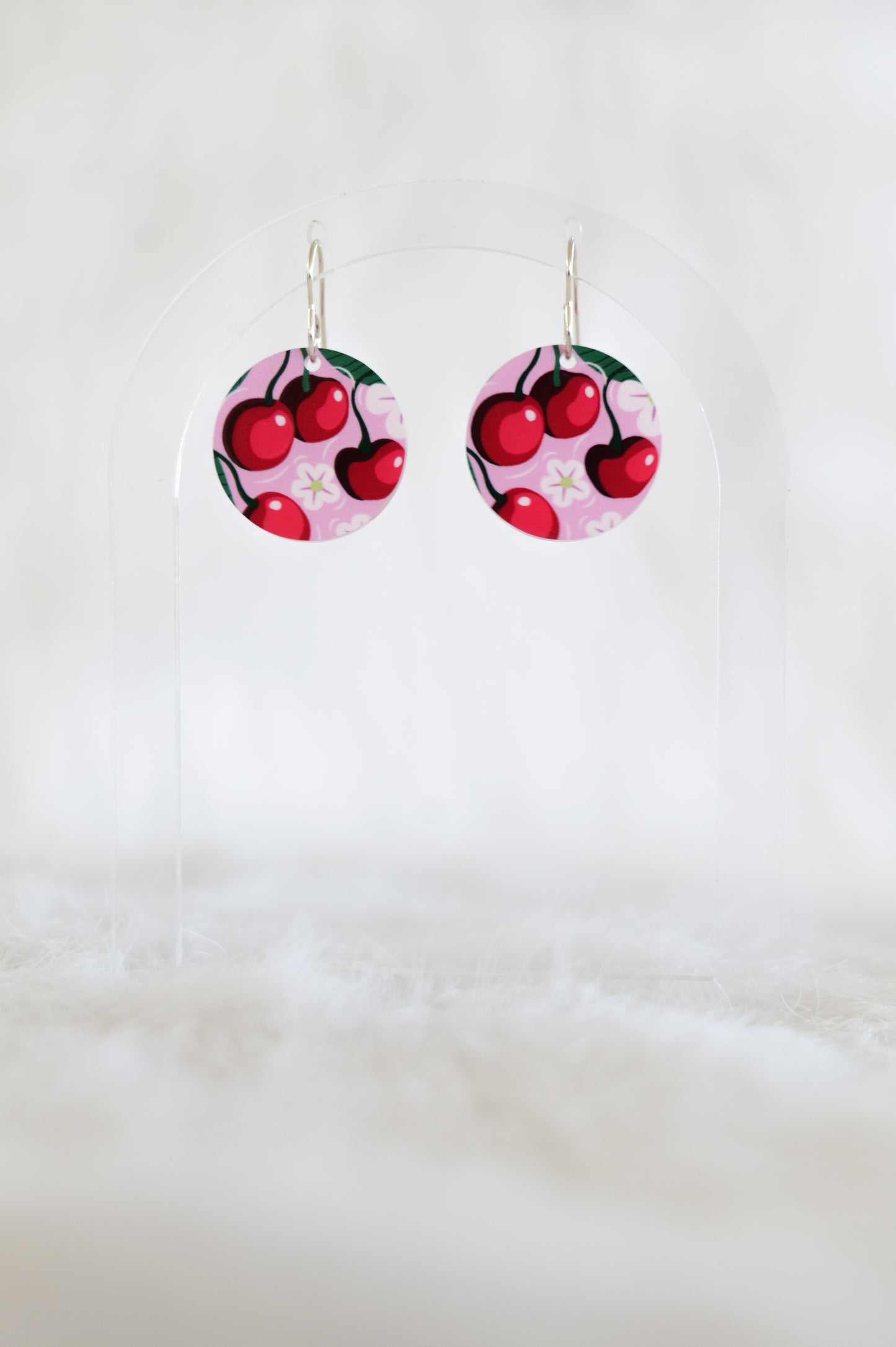 Pair of round earrings showing red cherries and white flowers on a pink background, displayed on a clear stand against a white backdrop