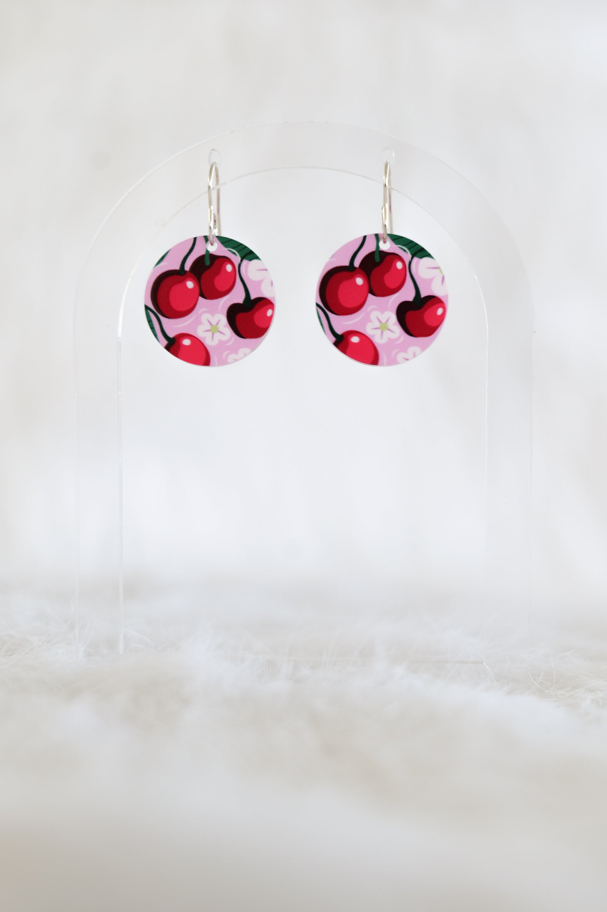 Pair of round earrings showing red cherries and white flowers on a pink background, displayed on a clear stand against a white backdrop