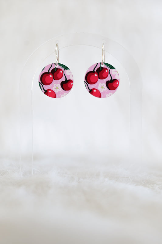 Pair of round earrings showing red cherries and white flowers on a pink background, displayed on a clear stand against a white backdrop