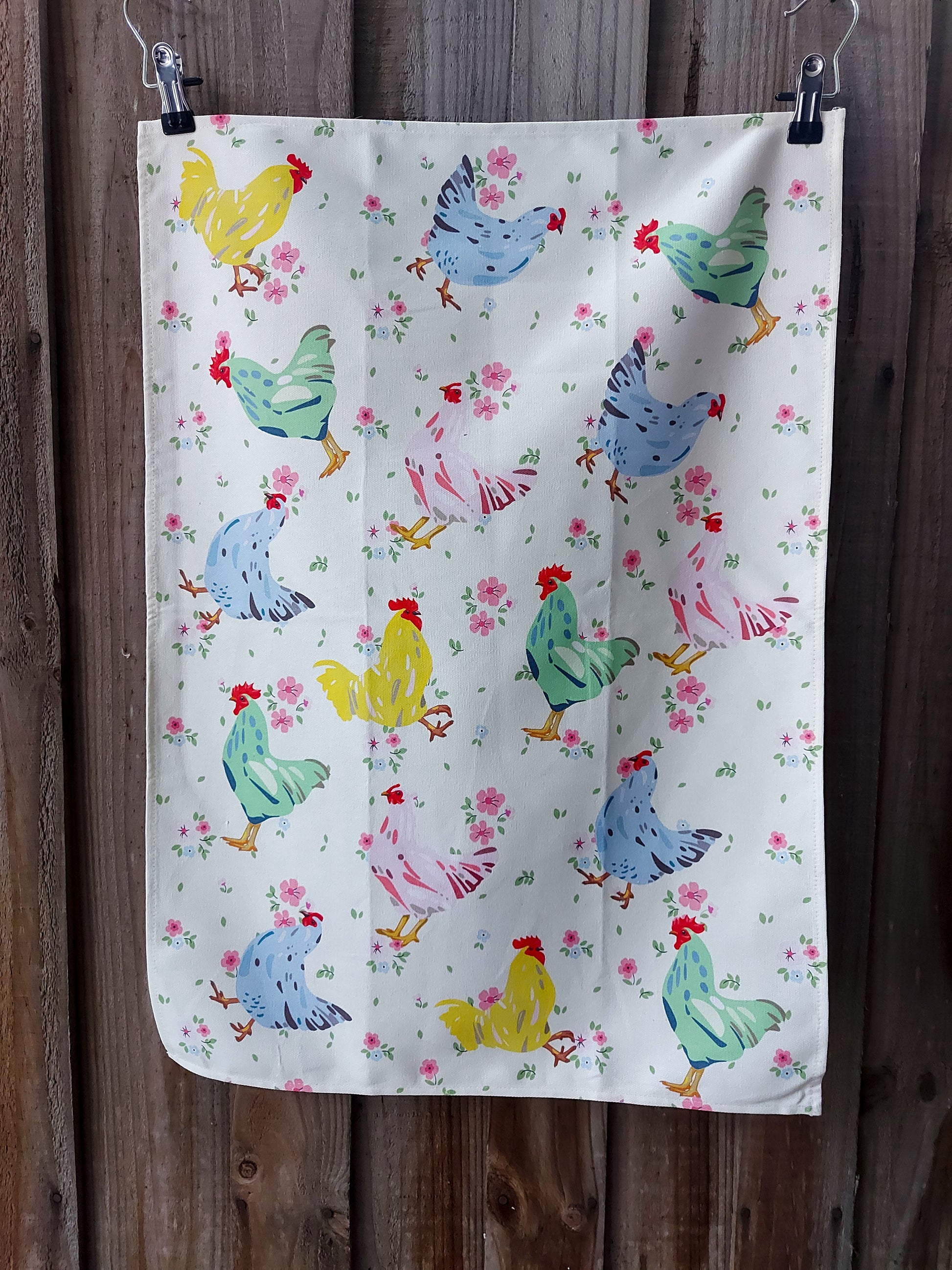 Cotton tea towel featuring chooks in pink, blue, green and yellow on a cream background with small pink flowers; displayed against a wooden fence