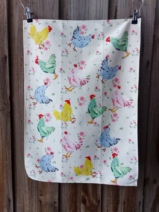 Cotton tea towel featuring chooks in pink, blue, green and yellow on a cream background with small pink flowers; displayed against a wooden fence