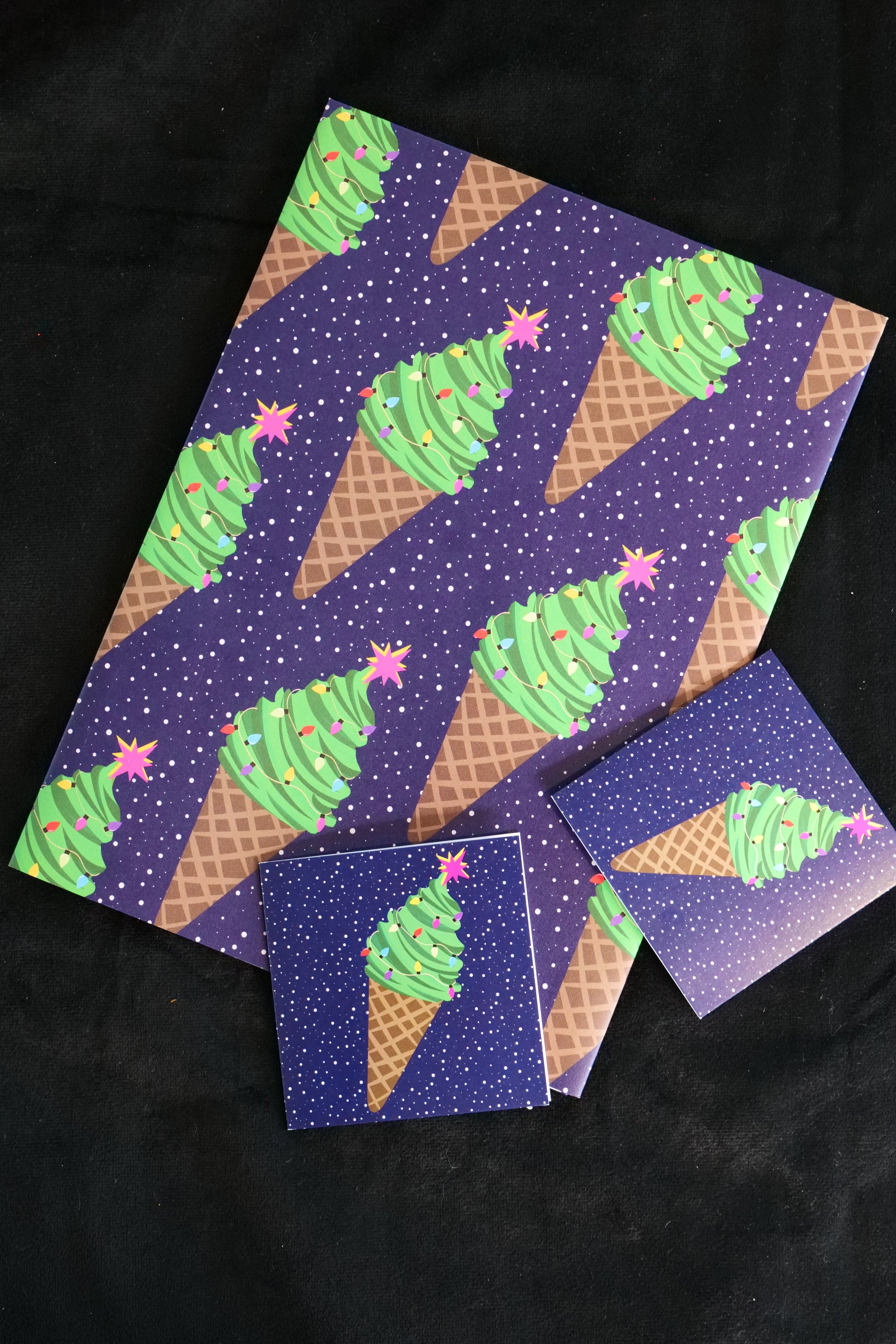 A folded sheet of gift wrap with ice cream cones that look like Christmas trees with stars on the top, set on a navy speckled background, shown against a black backdrop