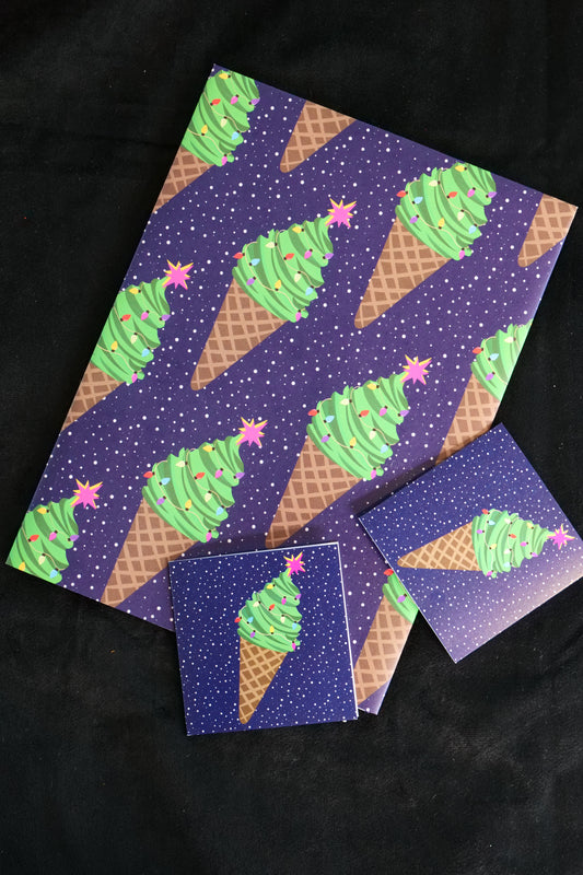 A folded sheet of gift wrap with ice cream cones that look like Christmas trees with stars on the top, set on a navy speckled background, shown against a black backdrop