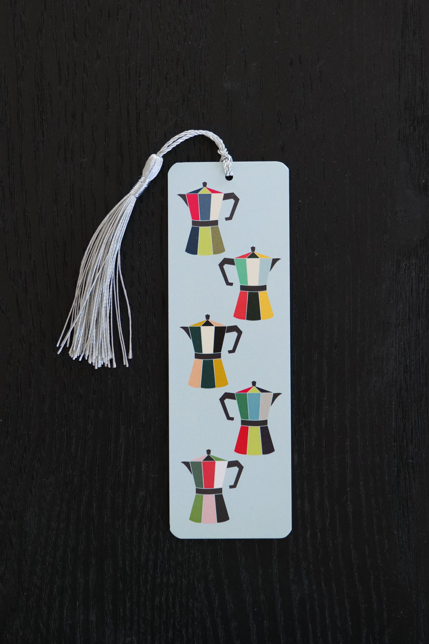 Bookmark featuring five brightly coloured coffee pots on a pale blue background with an ice blue tassel, sitting against a black backdrop