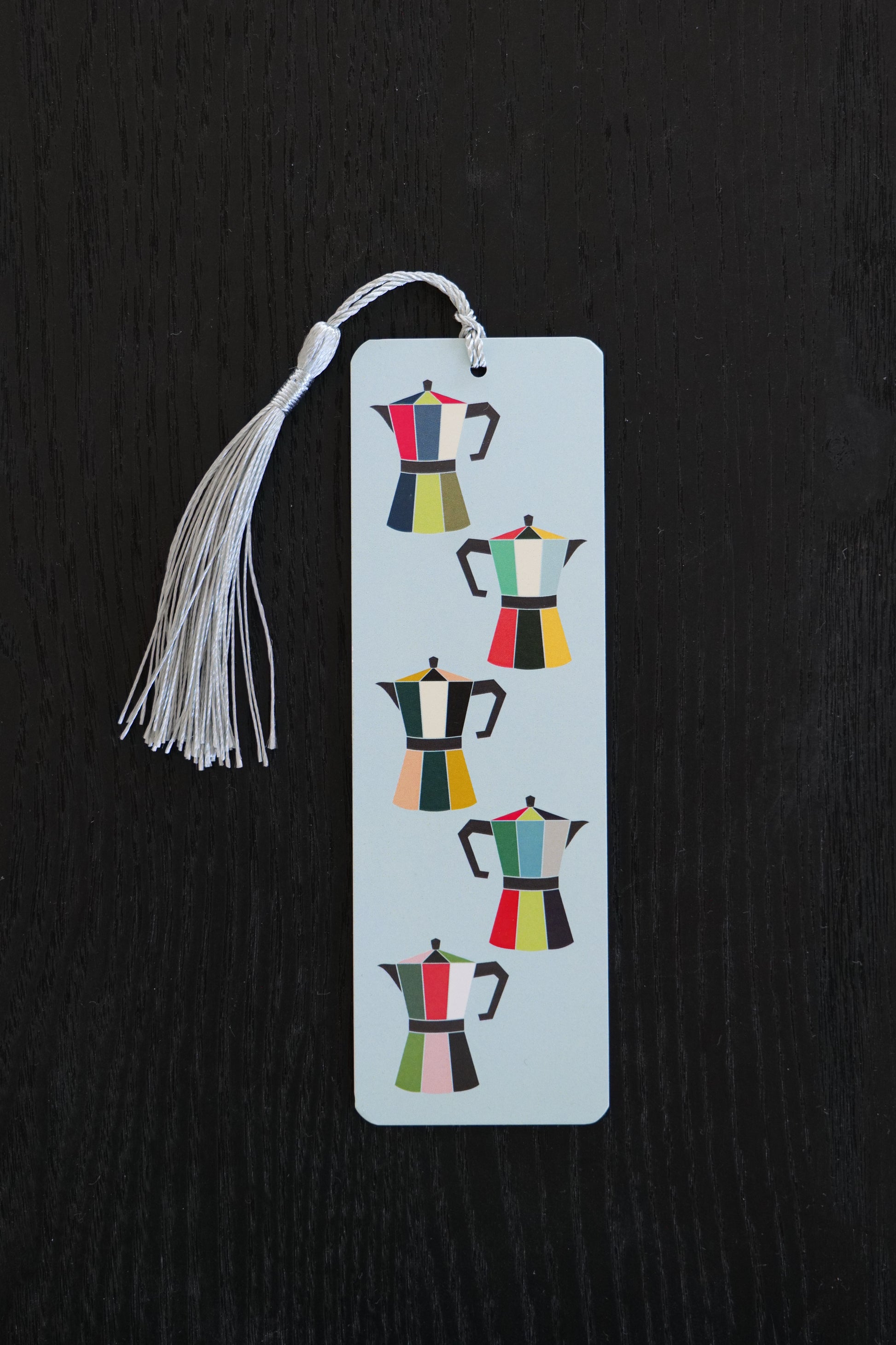 Bookmark featuring five brightly coloured coffee pots on a pale blue background with an ice blue tassel, sitting against a black backdrop