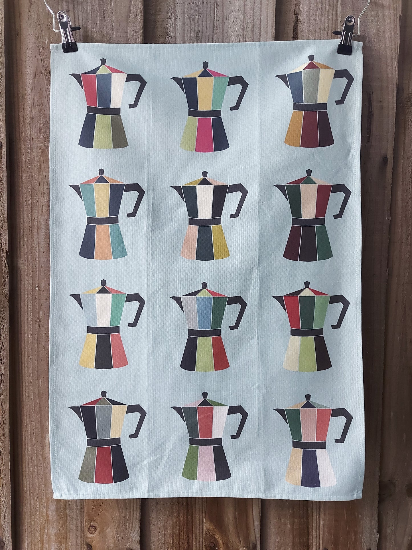 Light blue 100% cotton tea towel featuring a grid of colourful retro‑style moka pot illustrations displayed against a wooden background; designed, printed and made in Australia.