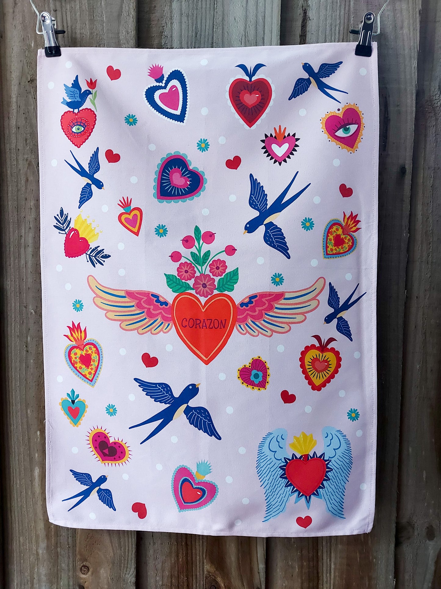 Cotton tea towel featuring a folk‑art inspired pattern of stylised hearts, winged motifs and blue birds on a pale pink background; displayed against a wooden fence