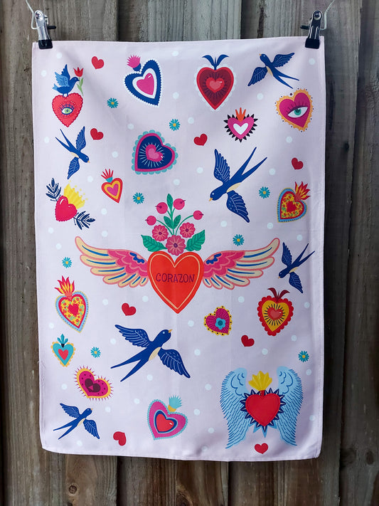Cotton tea towel featuring a folk‑art inspired pattern of stylised hearts, winged motifs and blue birds on a pale pink background; displayed against a wooden fence
