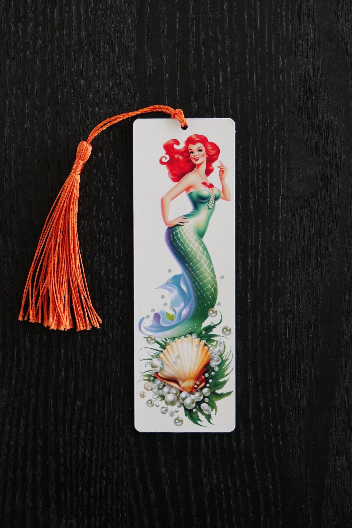 Bookmark featuring a green mermaid with red hair in 50s style with a scallop shell, seaweed and pearls on a cream background with an orange tassel, sitting against a black backdrop