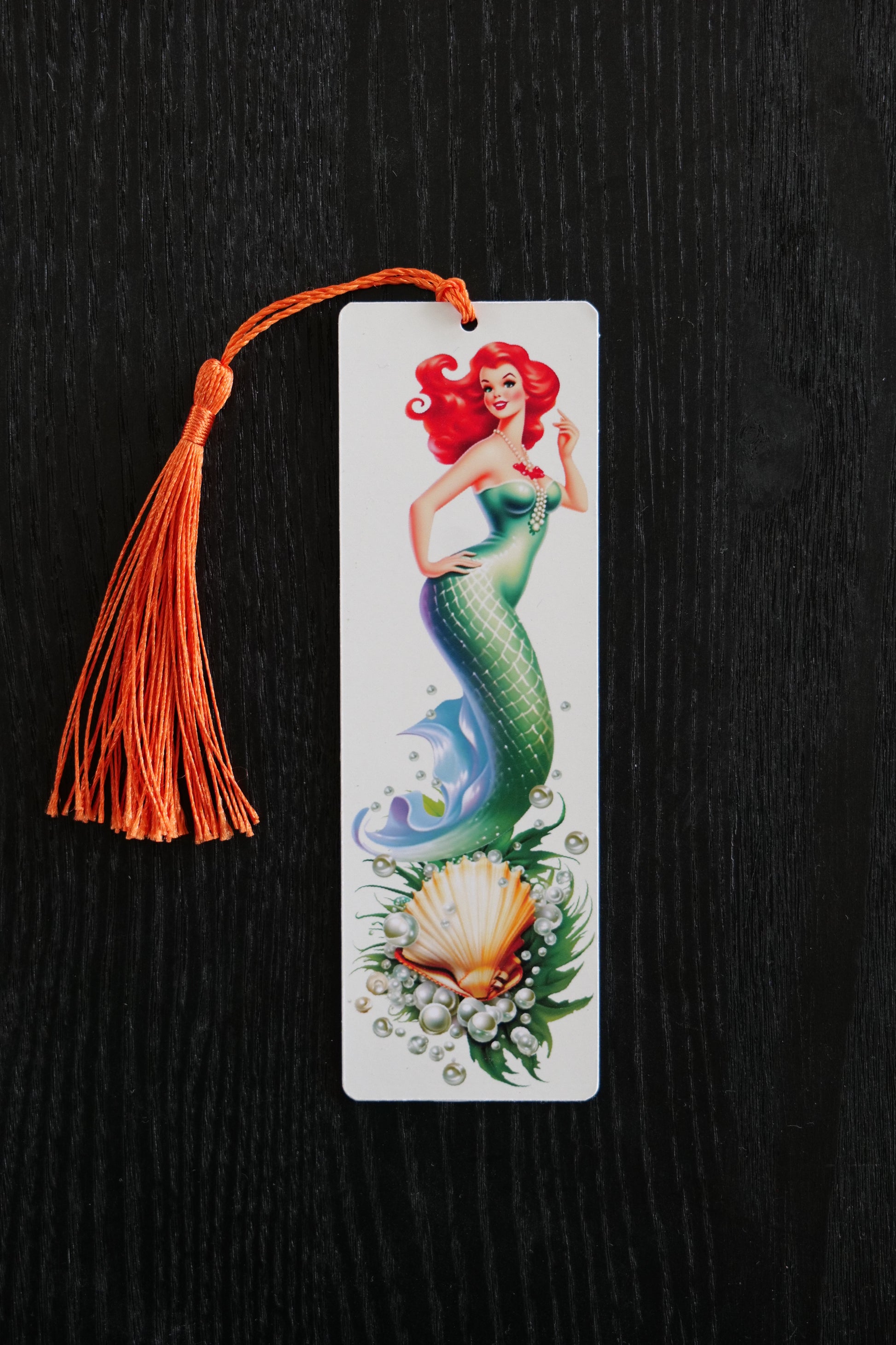 Bookmark featuring a green mermaid with red hair in 50s style with a scallop shell, seaweed and pearls on a cream background with an orange tassel, sitting against a black backdrop