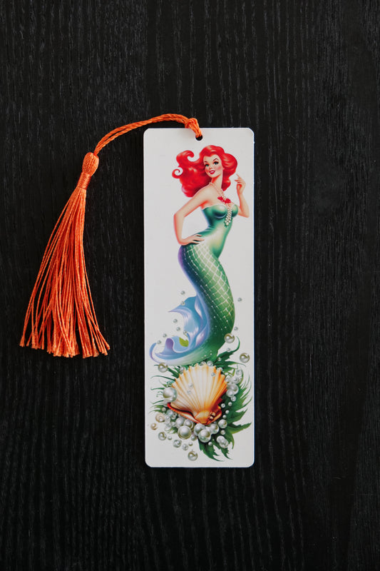 Bookmark featuring a green mermaid with red hair in 50s style with a scallop shell, seaweed and pearls on a cream background with an orange tassel, sitting against a black backdrop