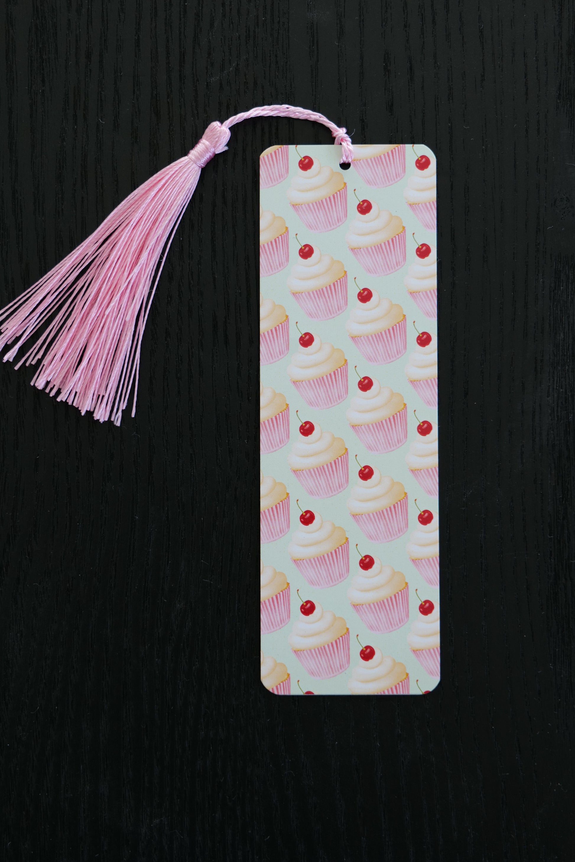 Bookmark featuring a repeating pattern of pale pink cupcakes with cherries on top with a light green background and a pale pink tassel, sitting against a black backdrop