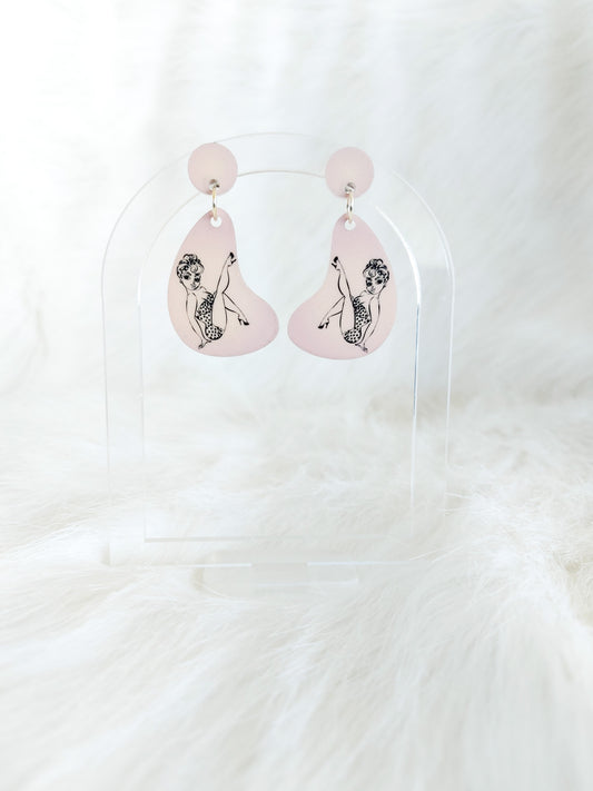 Pale pink boomerang shaped earrings with black pin-up style line drawings of a girl, displayed on a clear stand against a white background